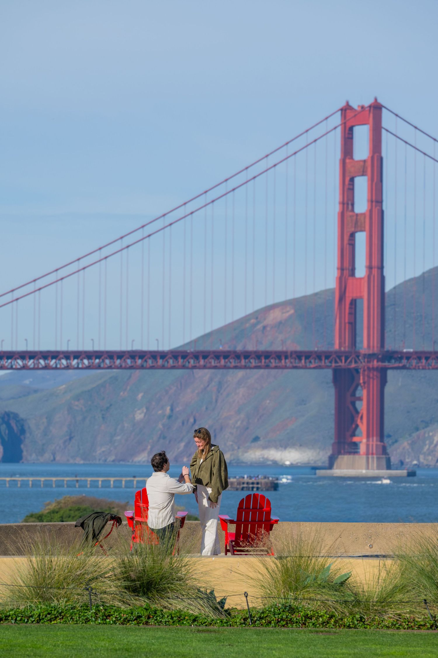Two people chat near red Adirondack chairs with the iconic Golden Gate Bridge and San Francisco Bay in the background.