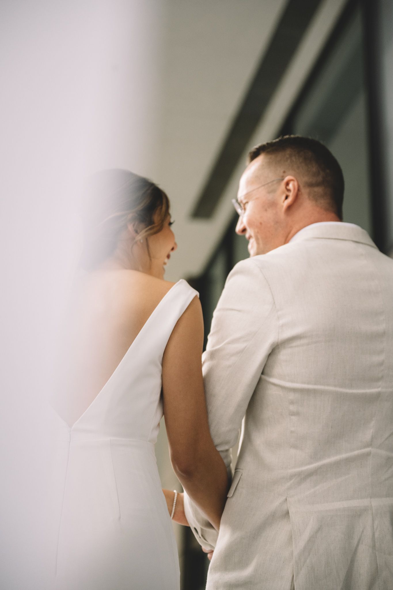 Bride in white gown smiles while holding hands with man in linen suit near large windows.