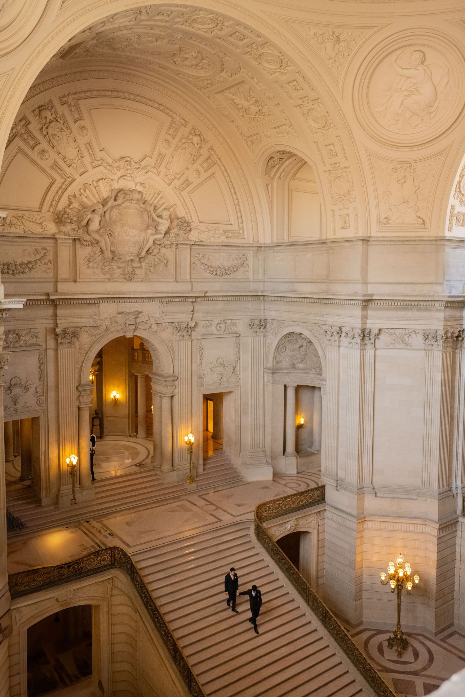 Aerial view of couple descending grand ornate marble staircase inside San Francisco City Hall.