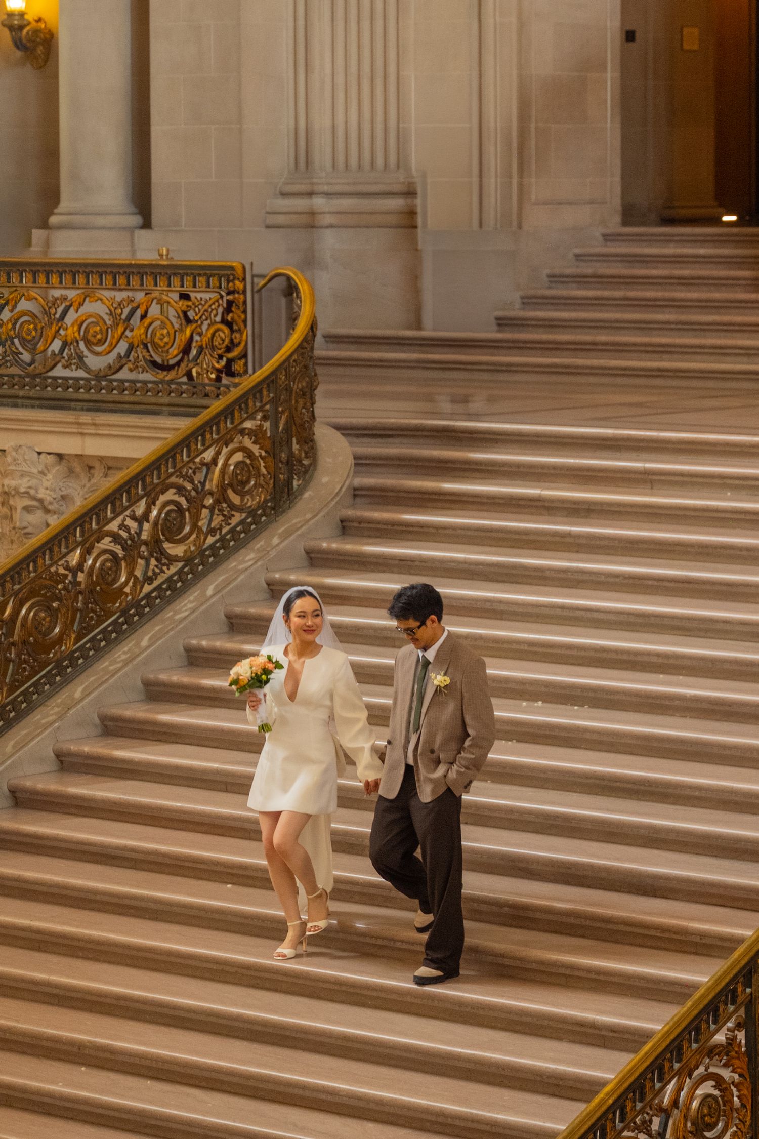 Bride in white mini dress and groom descend ornate golden staircase at city hall.