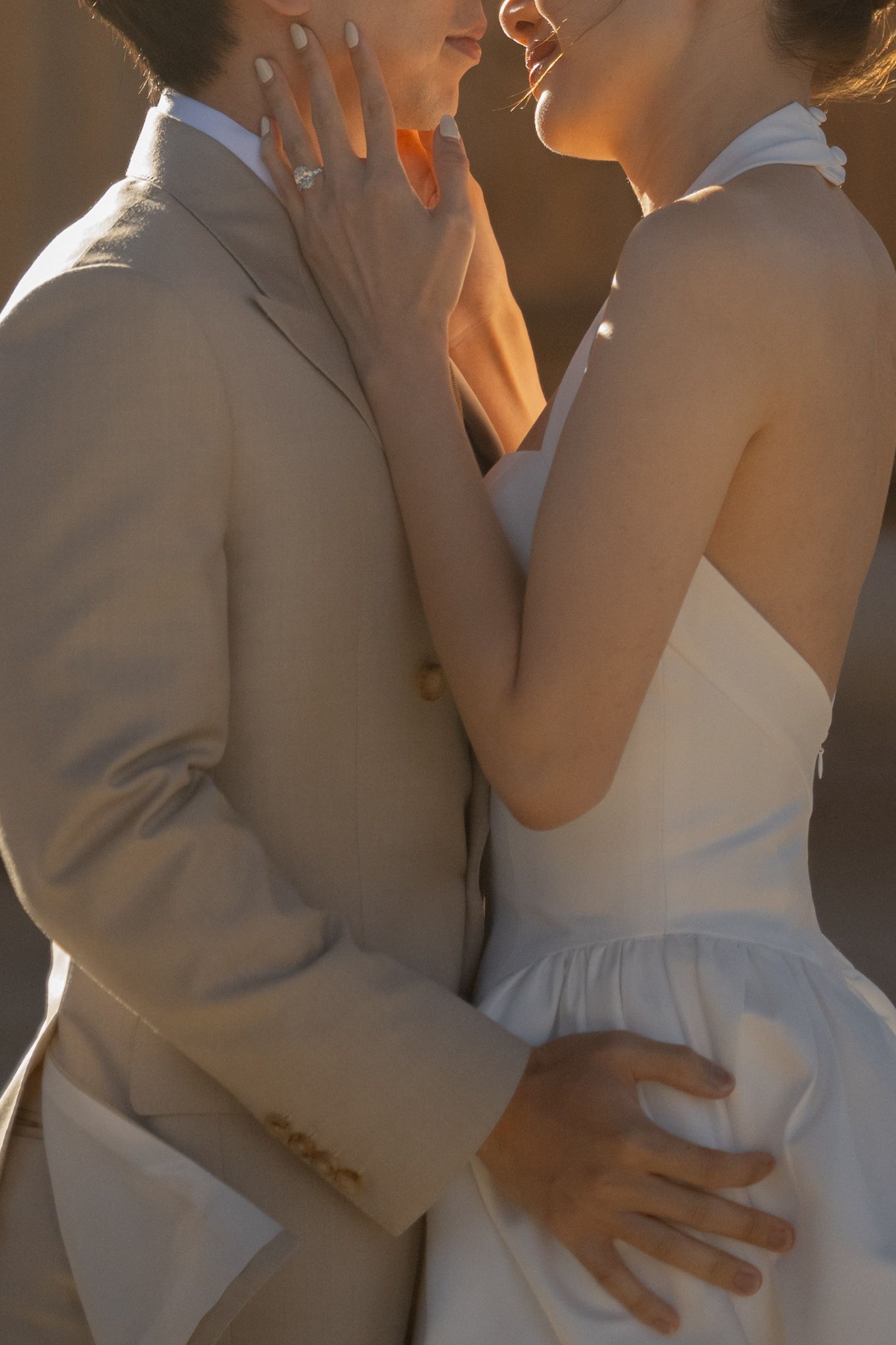 Close-up of a wedding couple embracing, bride in white halter dress and groom in beige suit, golden sunset light.