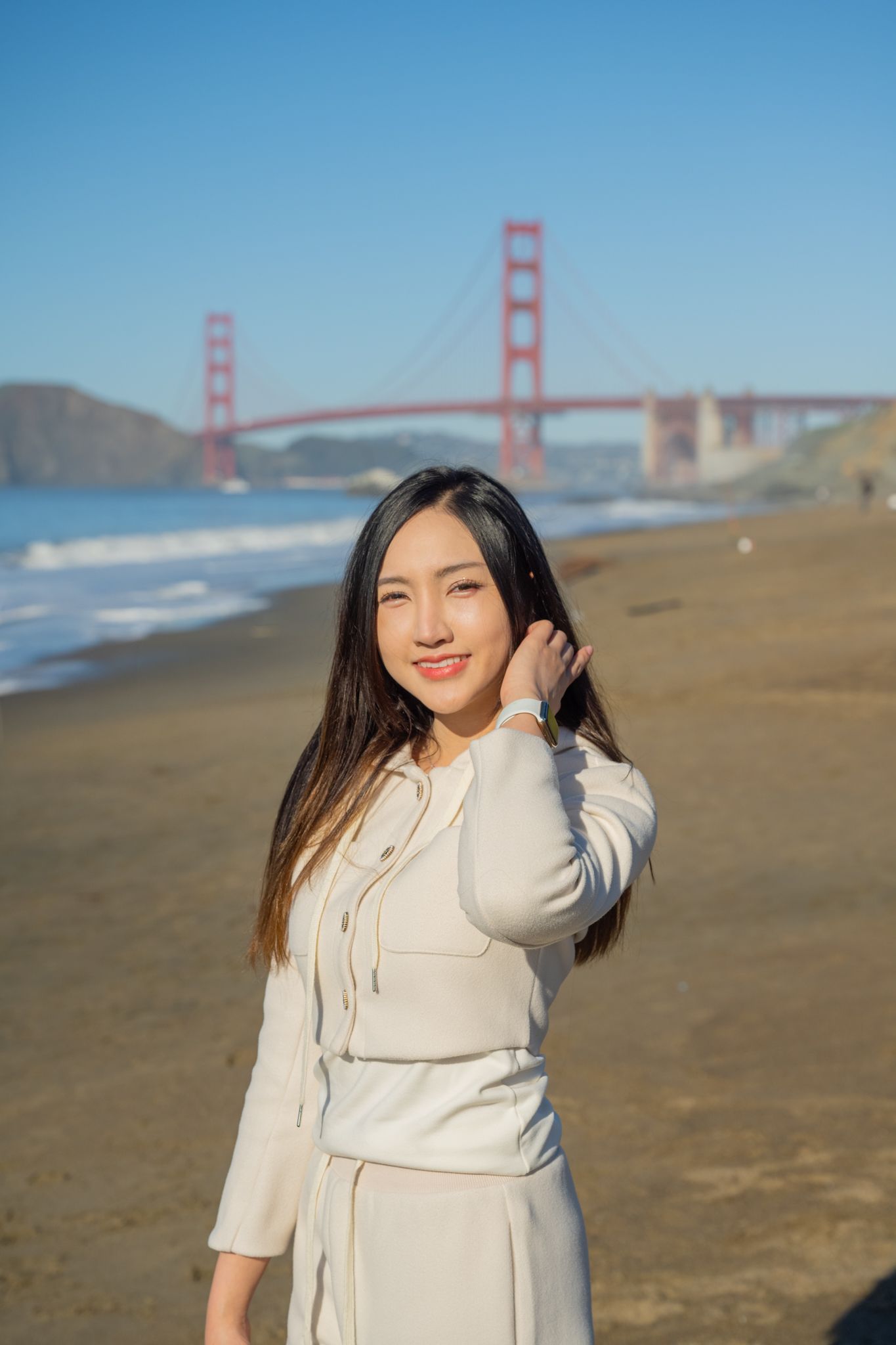 Smiling young woman in cream outfit stands on beach with Golden Gate Bridge in background.