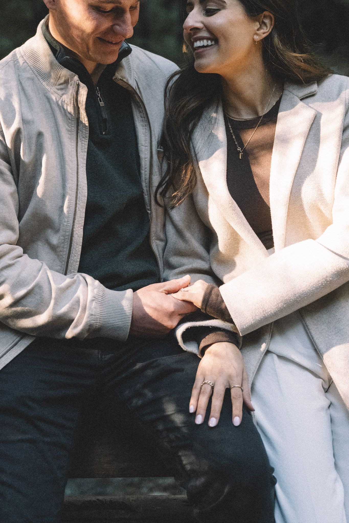 Smiling couple sits on bench holding hands, woman wearing engagement ring in a white blazer.