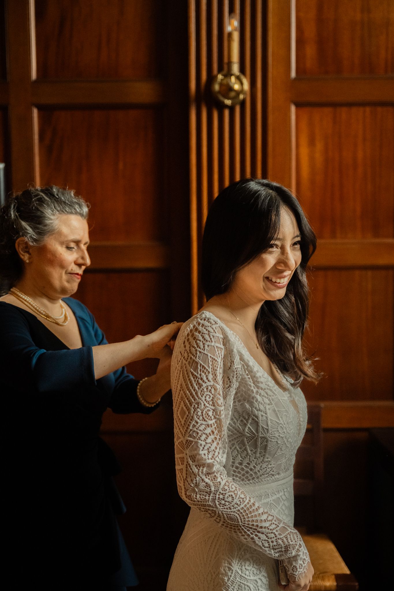 Older woman helps smiling Asian bride in lace wedding dress prepare in warm wood-paneled room.