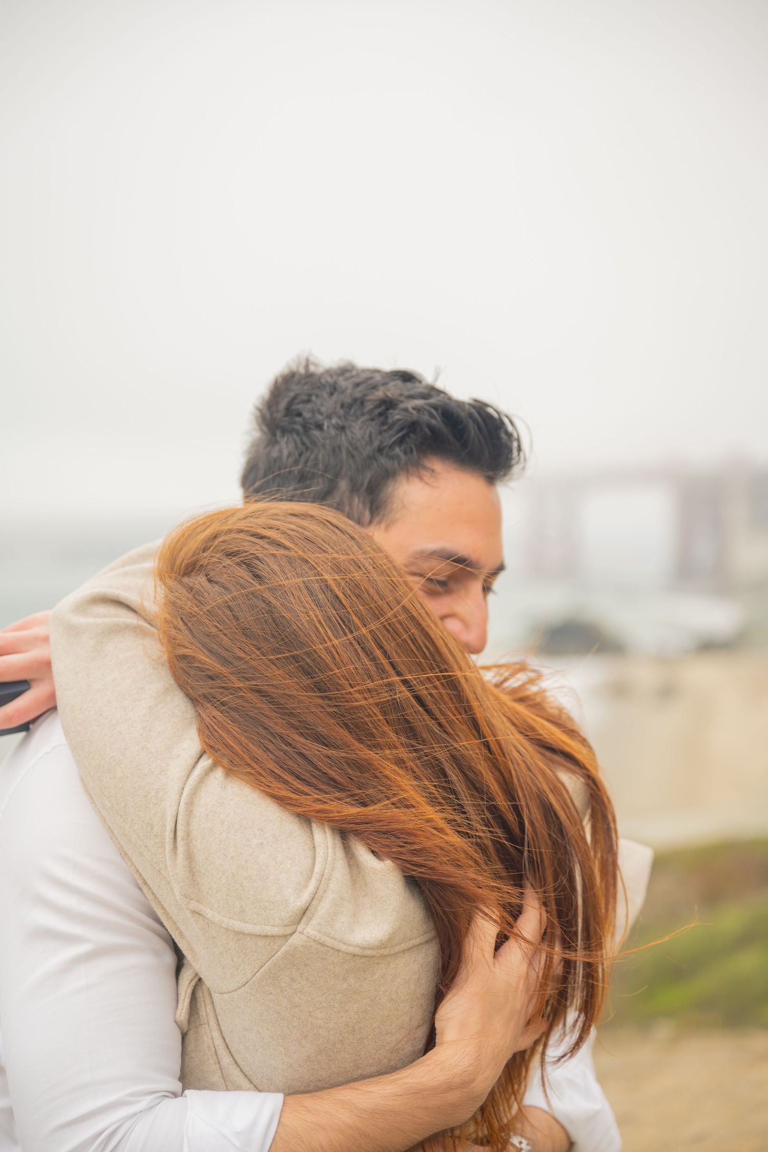 Couple shares tender windswept embrace outdoors with misty coastal cityscape in the background.