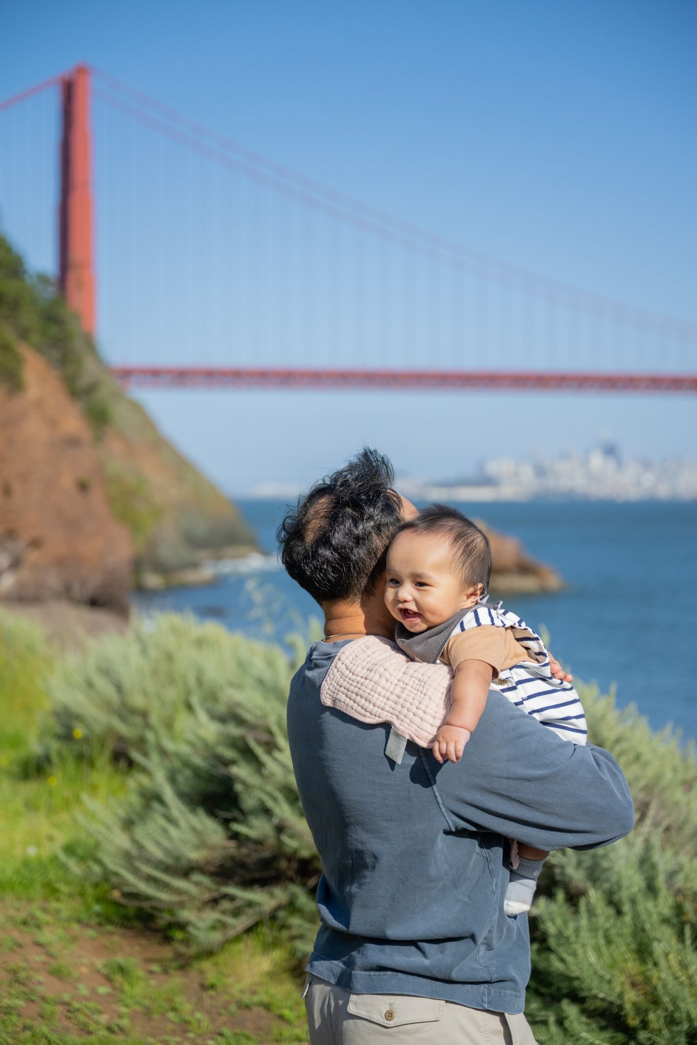 Parent holding baby near Golden Gate Bridge in San Francisco on a sunny day with blue skies and green shrubs.