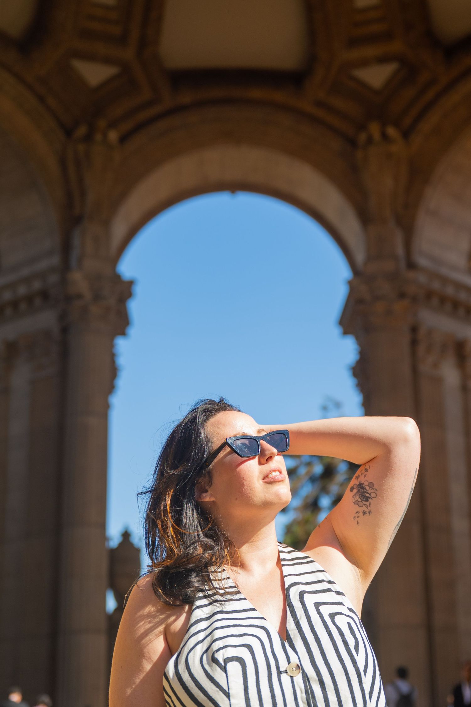 A woman in a striped dress and sunglasses poses under a grand stone arch on a sunny day.
