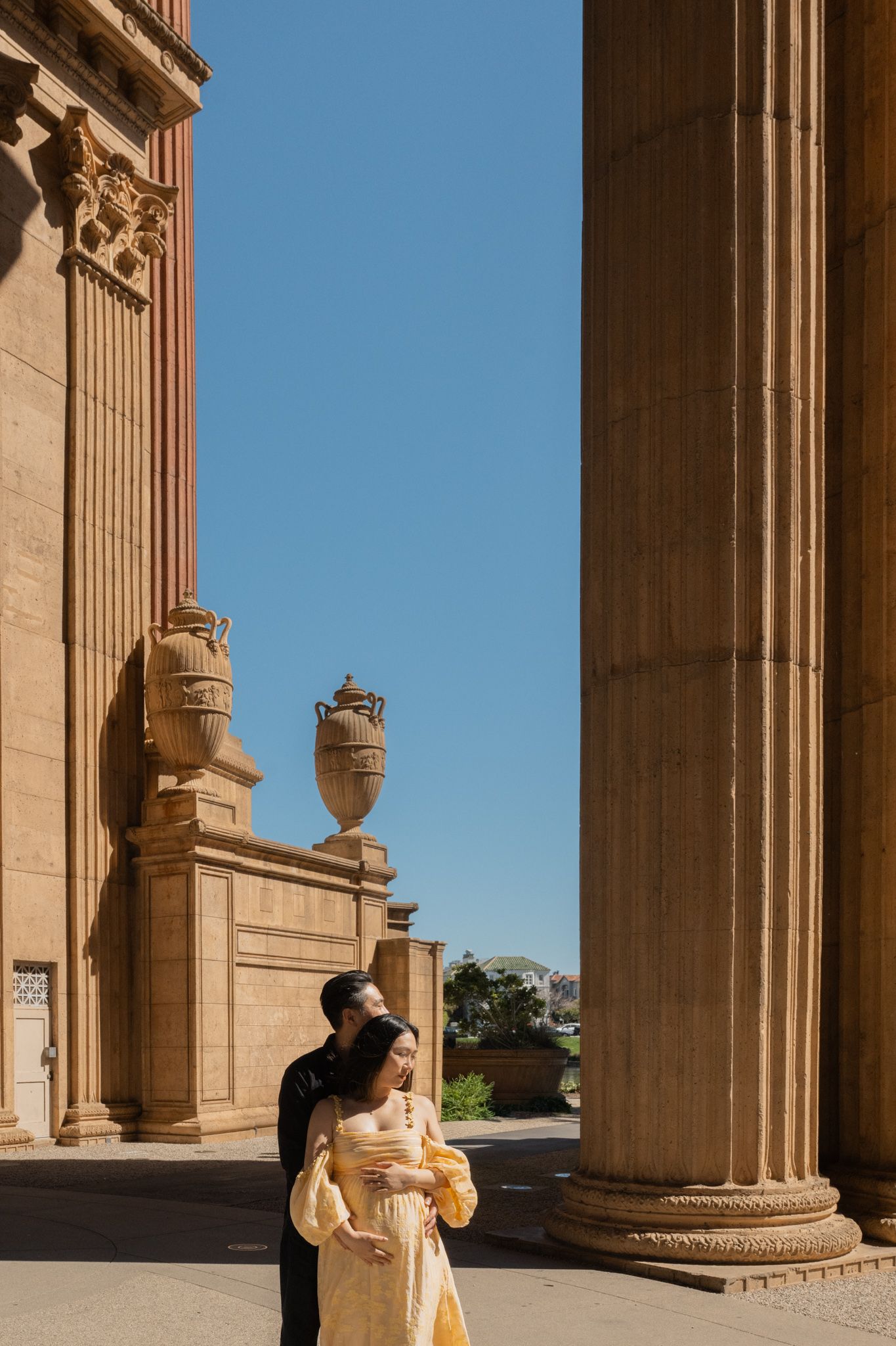 Couple embracing between ornate columns at the Palace of Fine Arts in San Francisco on a sunny day.