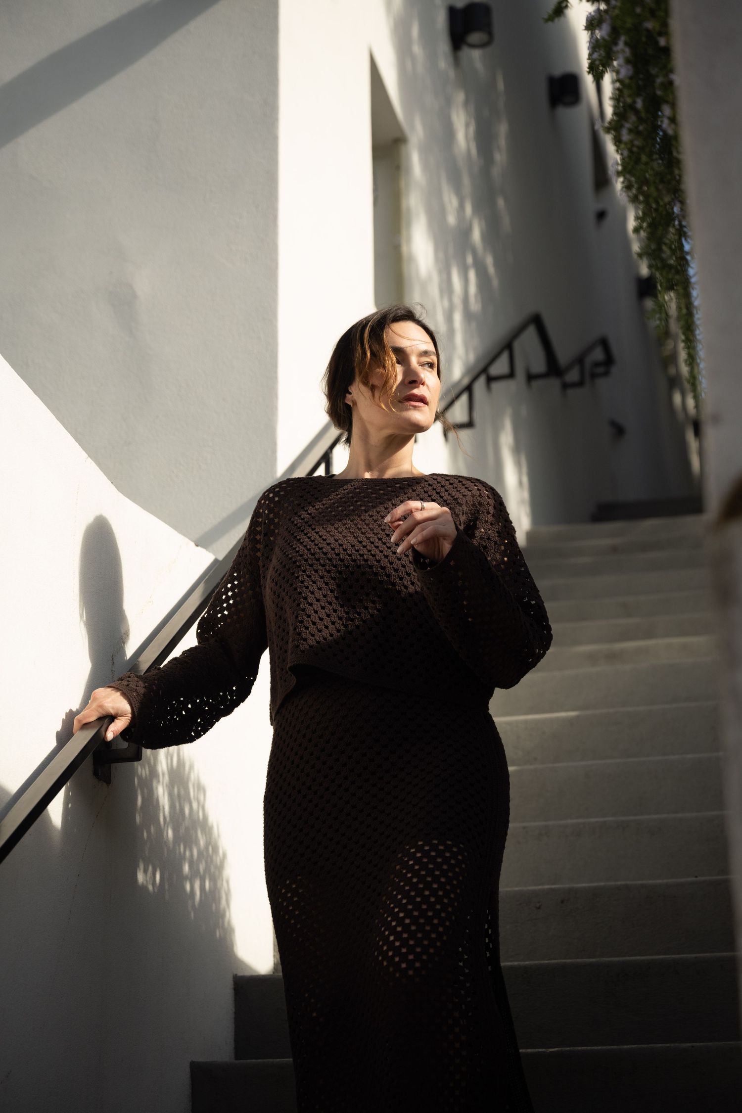 Woman in black mesh dress poses on sunlit white stairs, casting dramatic shadows on the wall.