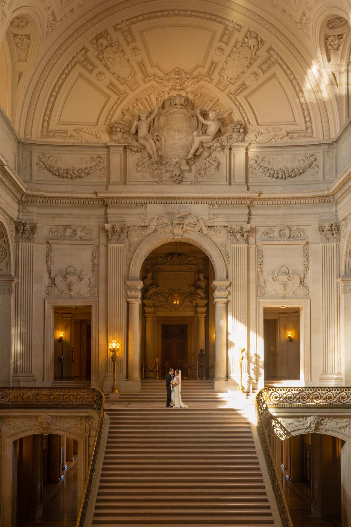Bride and groom embrace on grand staircase of San Francisco City Hall with ornate Beaux-Arts architecture.
