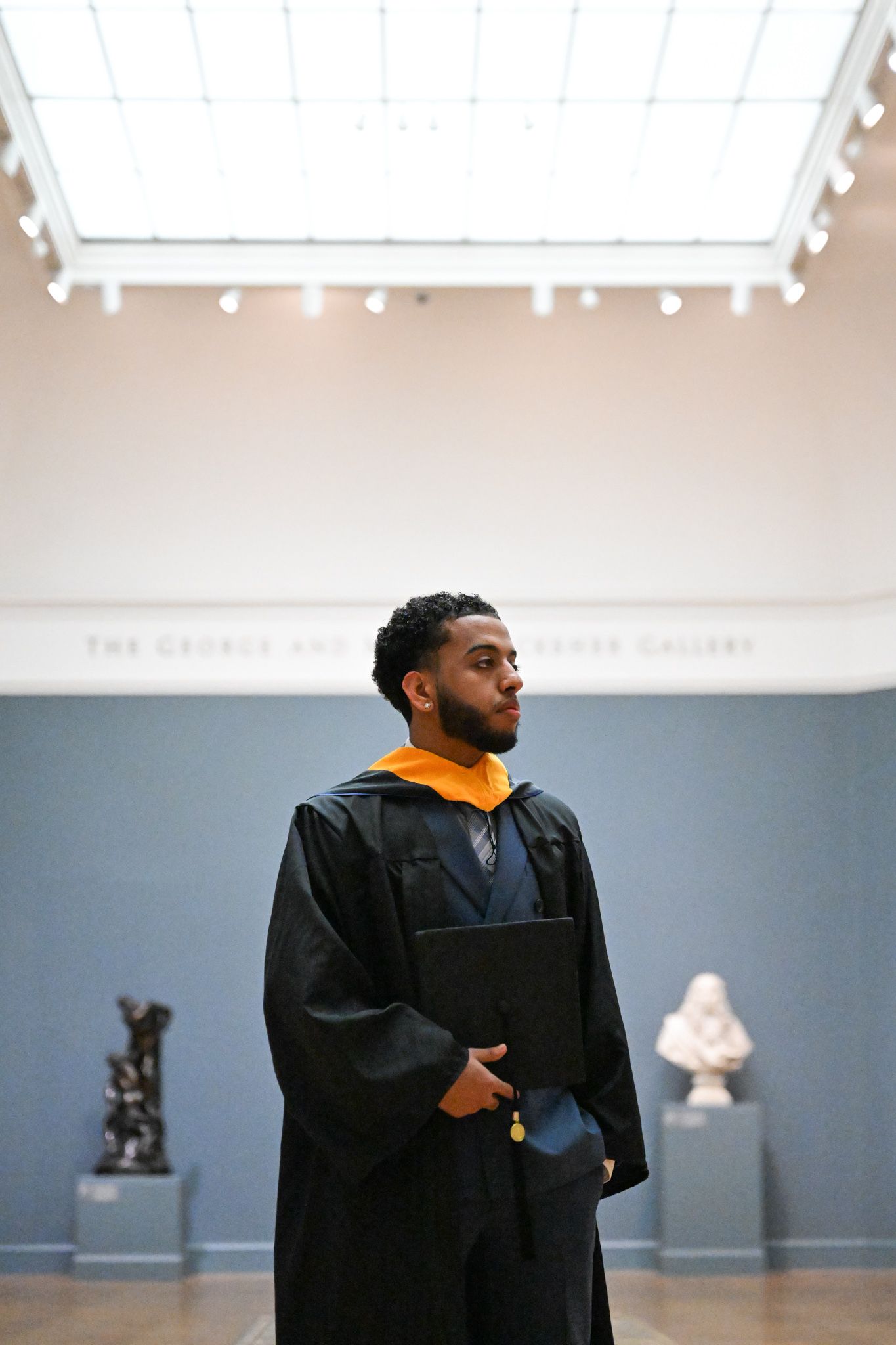 Graduate in black cap and gown holds diploma in a museum gallery with sculptures in background.