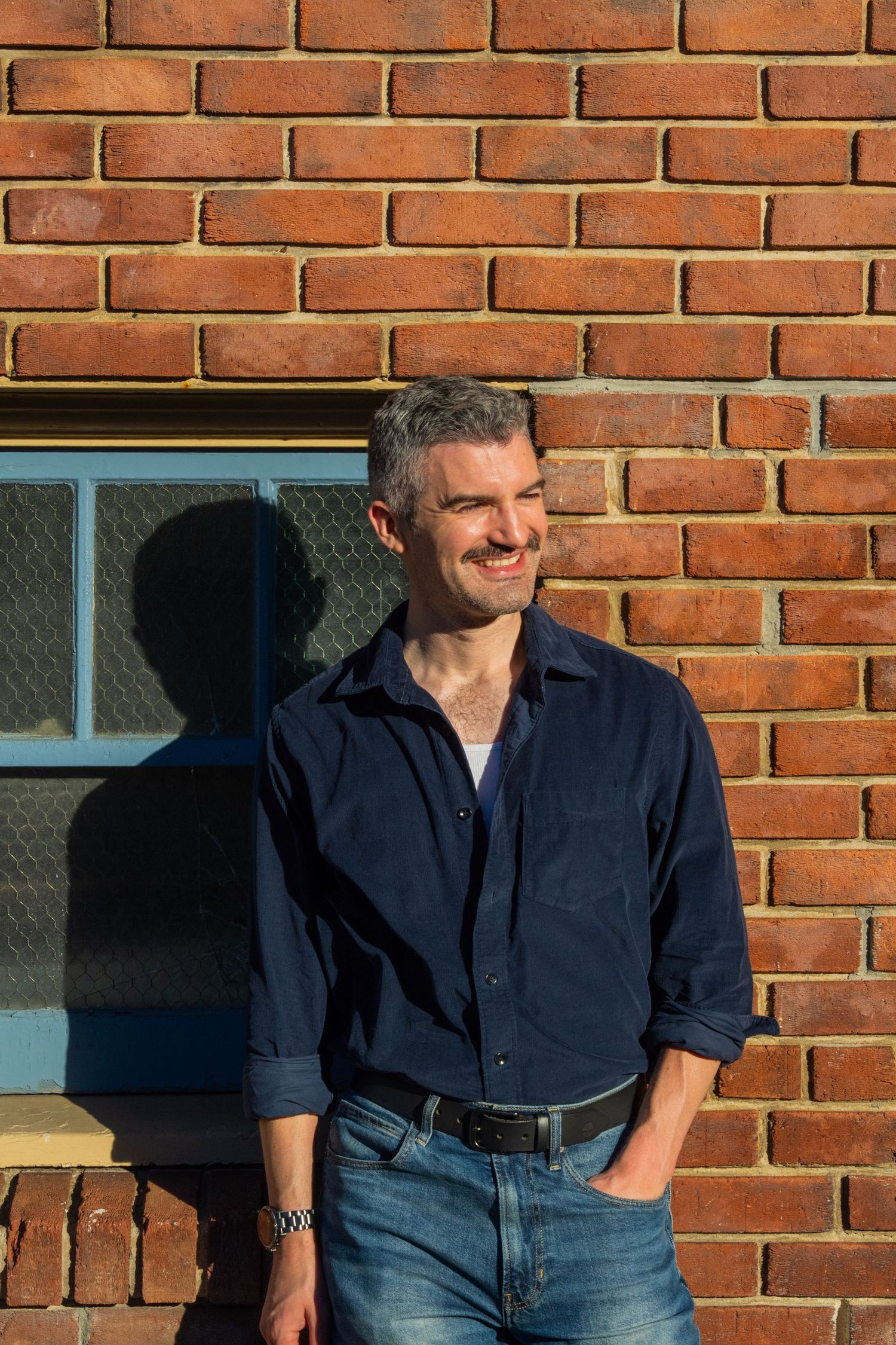A smiling middle-aged man in a navy shirt leans against a red brick wall in warm sunlight.