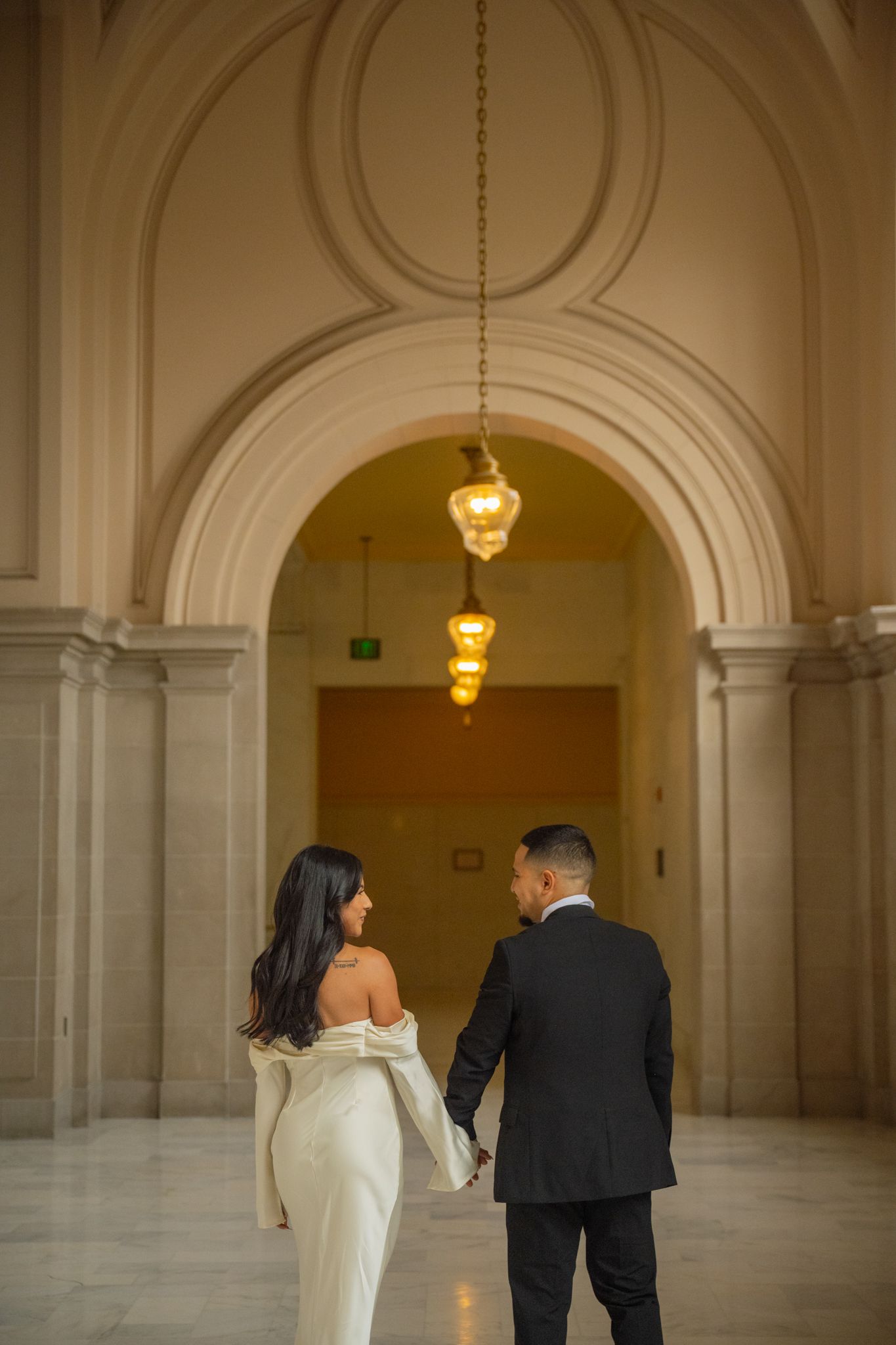 Wedding couple holds hands walking through an elegant arched corridor with warm pendant lighting.