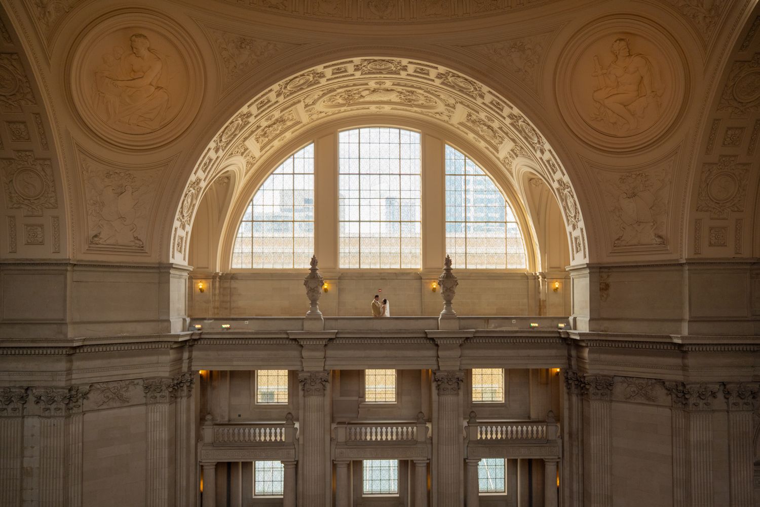 Wedding couple stands on balcony beneath grand arched window in a stunning beaux-arts building.