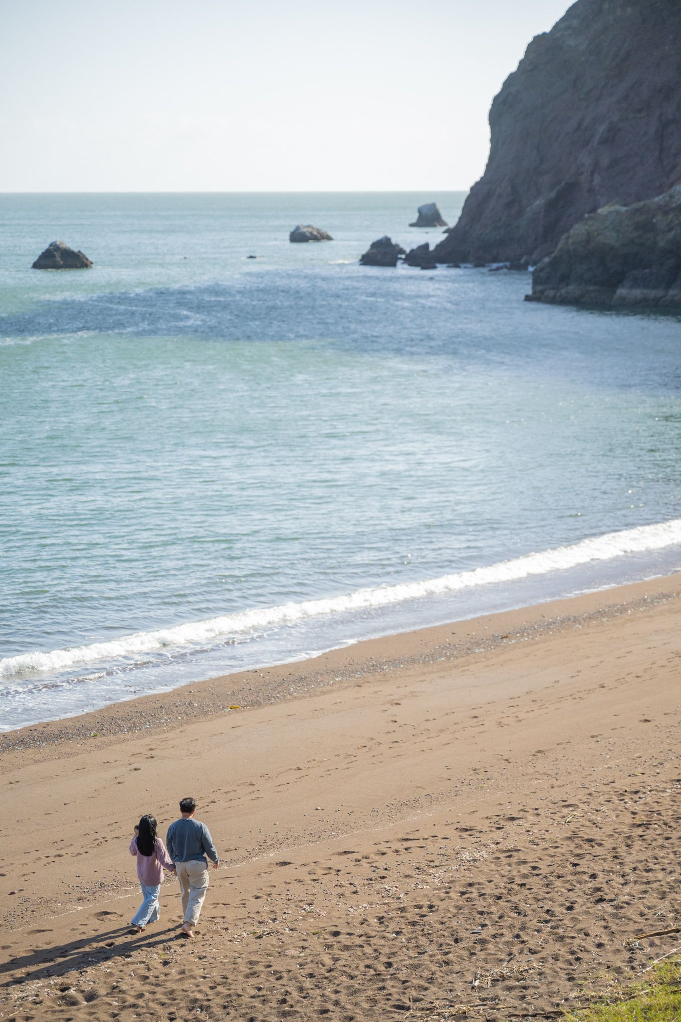 Couple walking hand in hand along a sandy beach with rocky cliffs and turquoise ocean waves in the background.