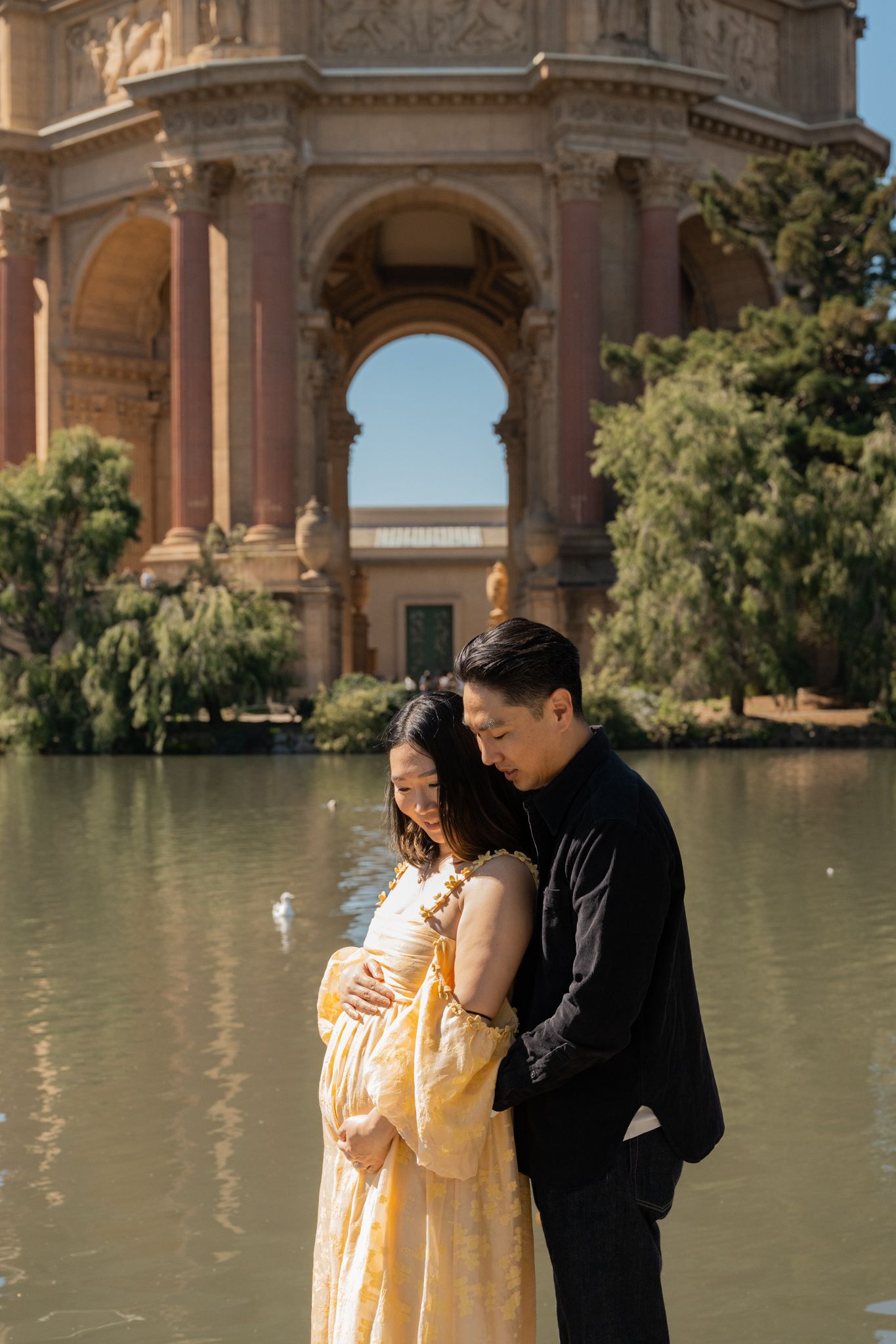 Couple tenderly embraces during maternity photo shoot at Palace of Fine Arts, San Francisco, with lagoon and rotunda backdrop.