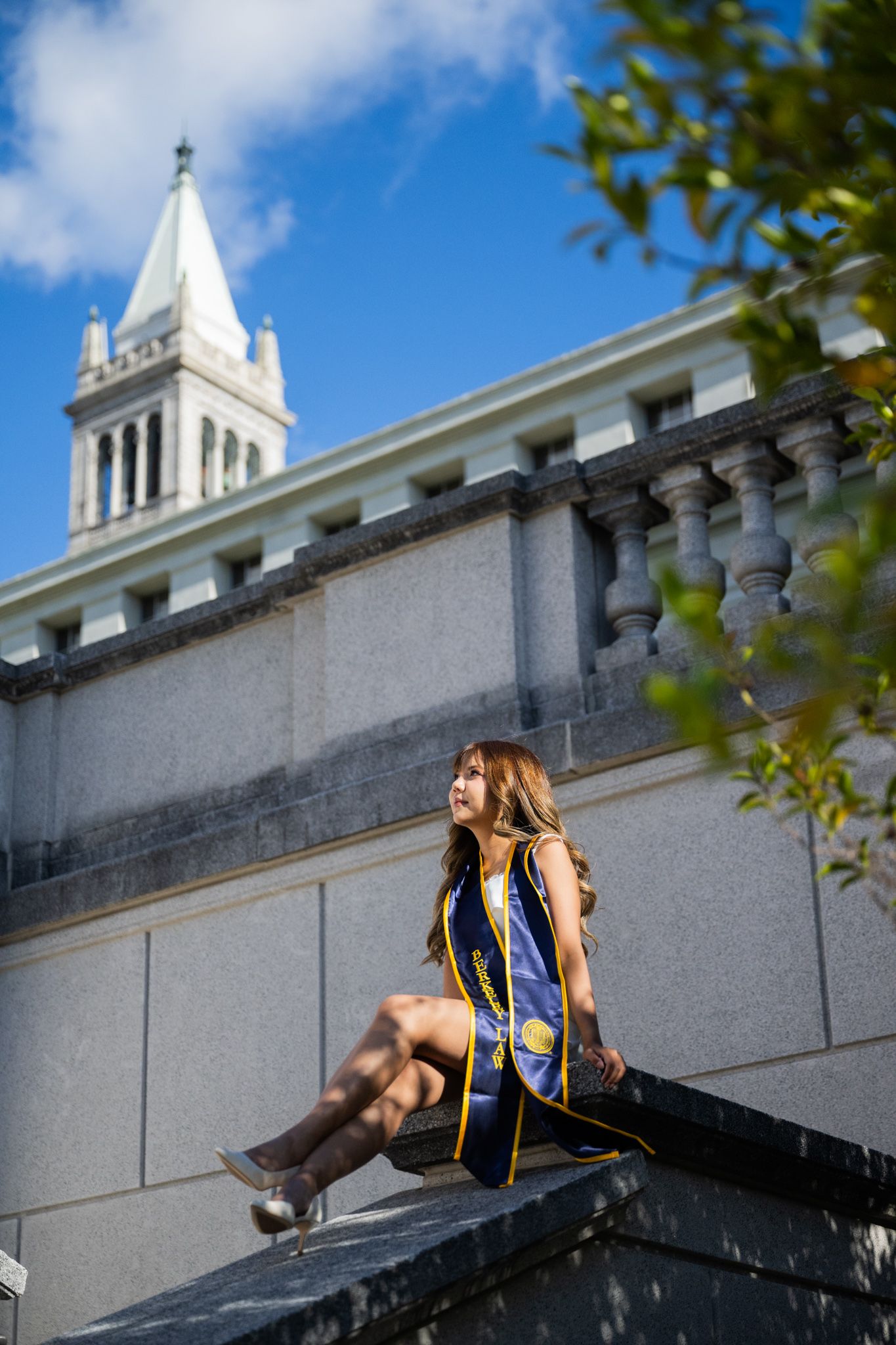 Female graduate in cap and gown sits on stone wall near a university bell tower on a sunny day.