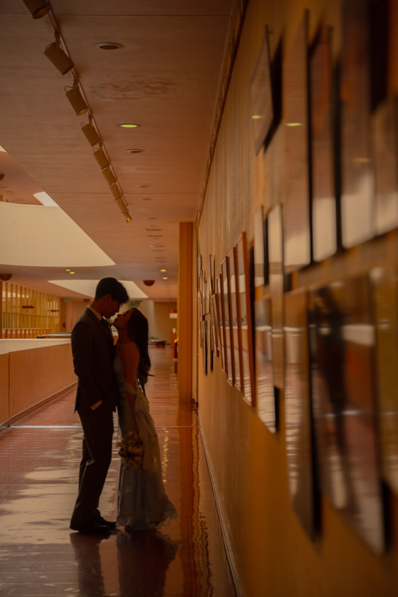 Bride and groom share an intimate moment in a dimly lit hallway lined with artwork.