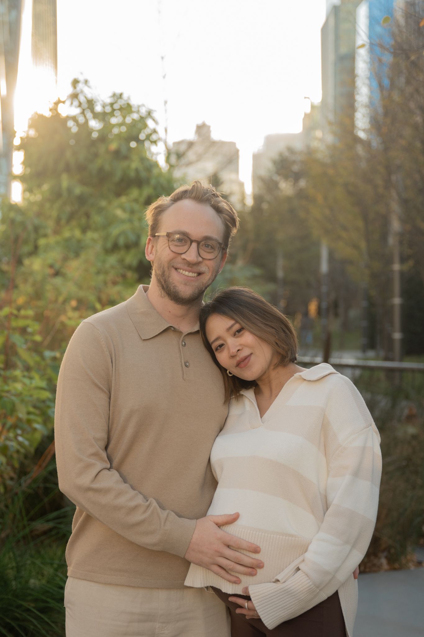 A smiling couple embraces outdoors at sunset, the woman visibly pregnant, with city buildings in the background.