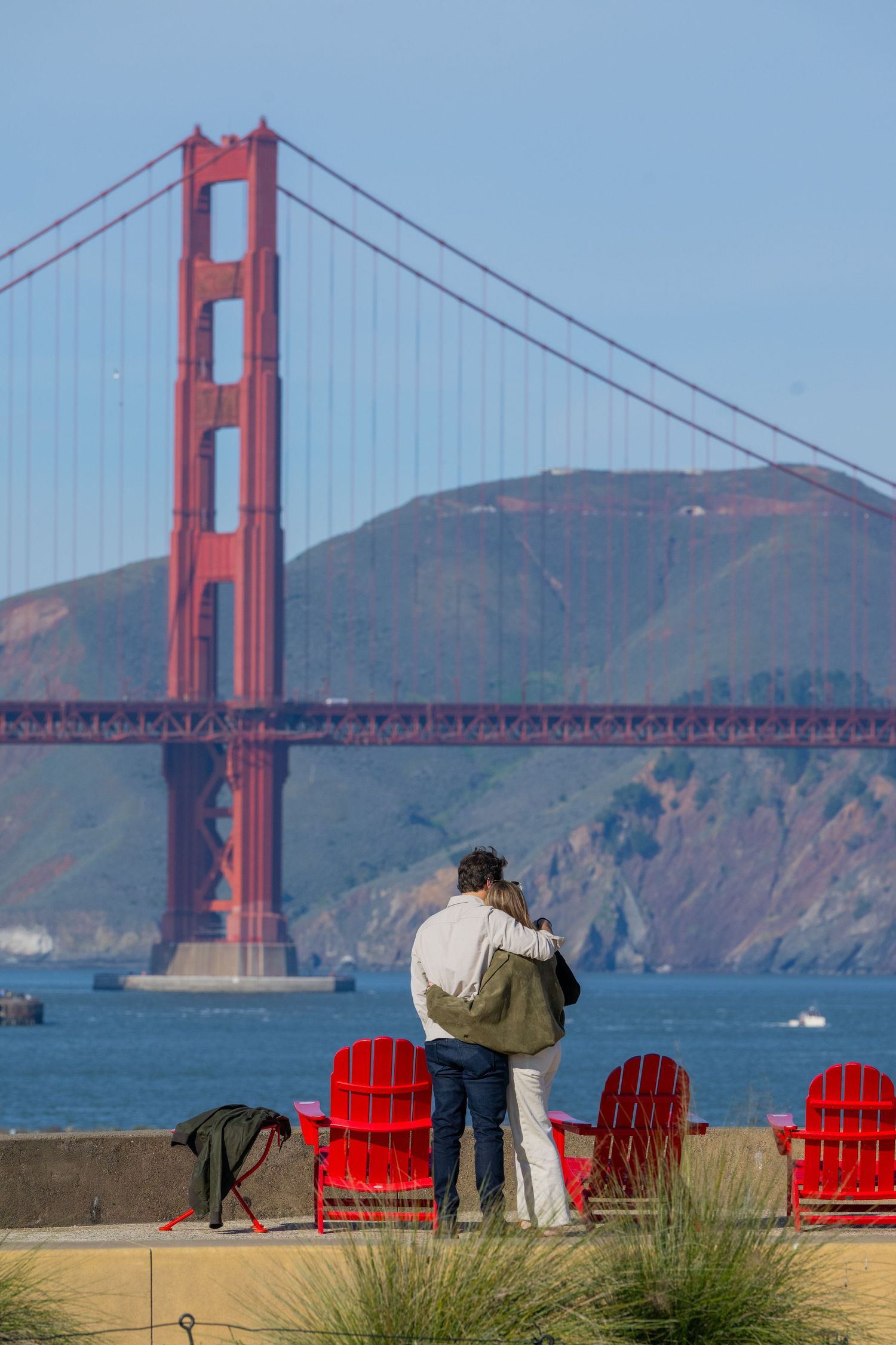 Couple embracing near red Adirondack chairs with iconic Golden Gate Bridge and Marin Headlands in background.