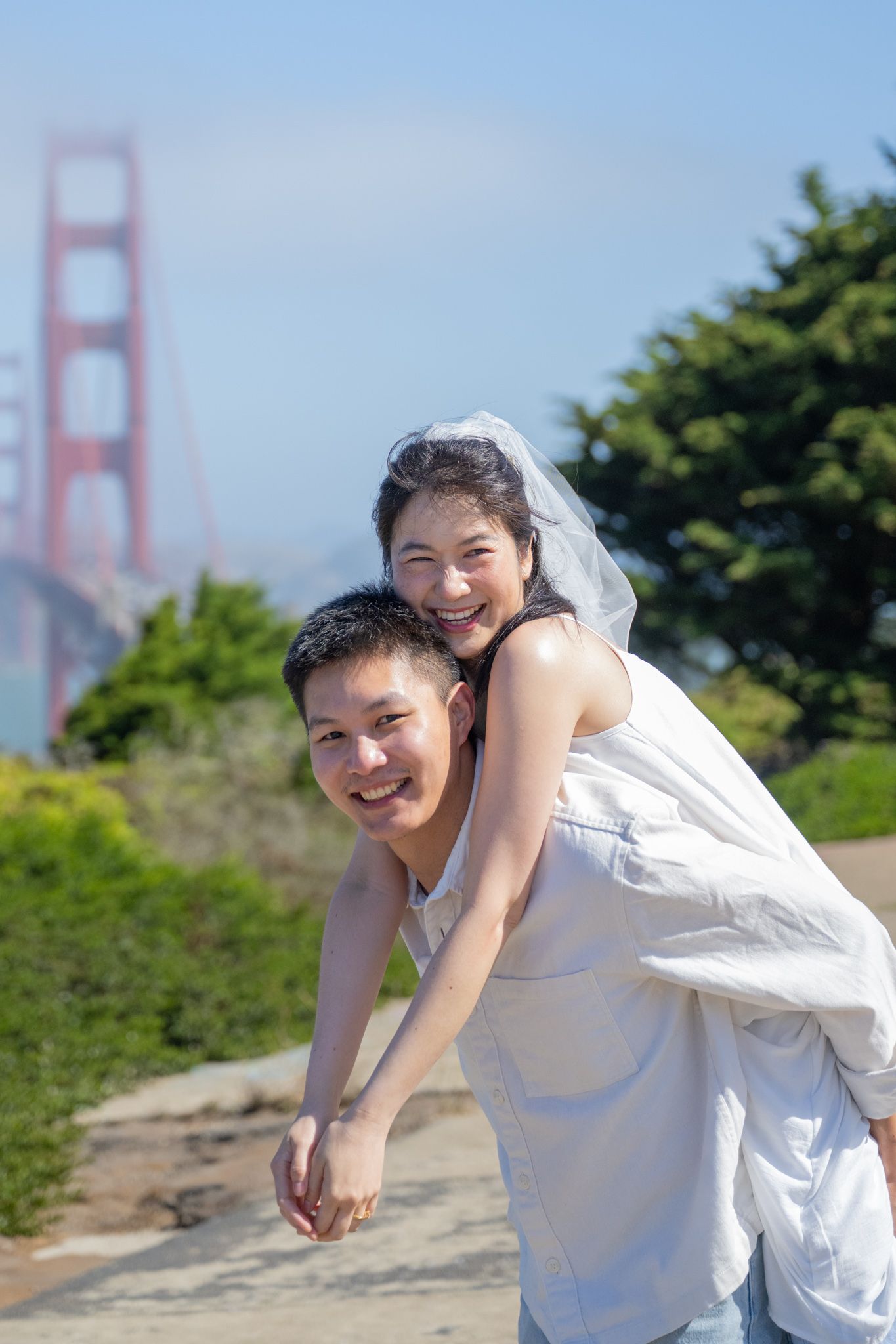 Asian couple in wedding attire laughing near Golden Gate Bridge, bride in white dress and veil on groom's back.