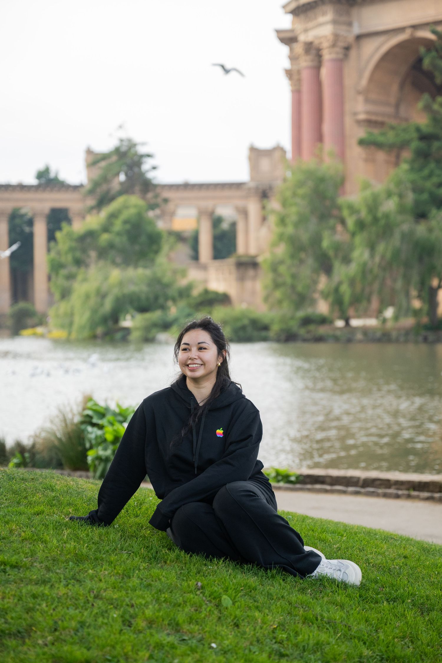Young woman in black Apple hoodie sits on grass smiling near the Palace of Fine Arts lagoon.