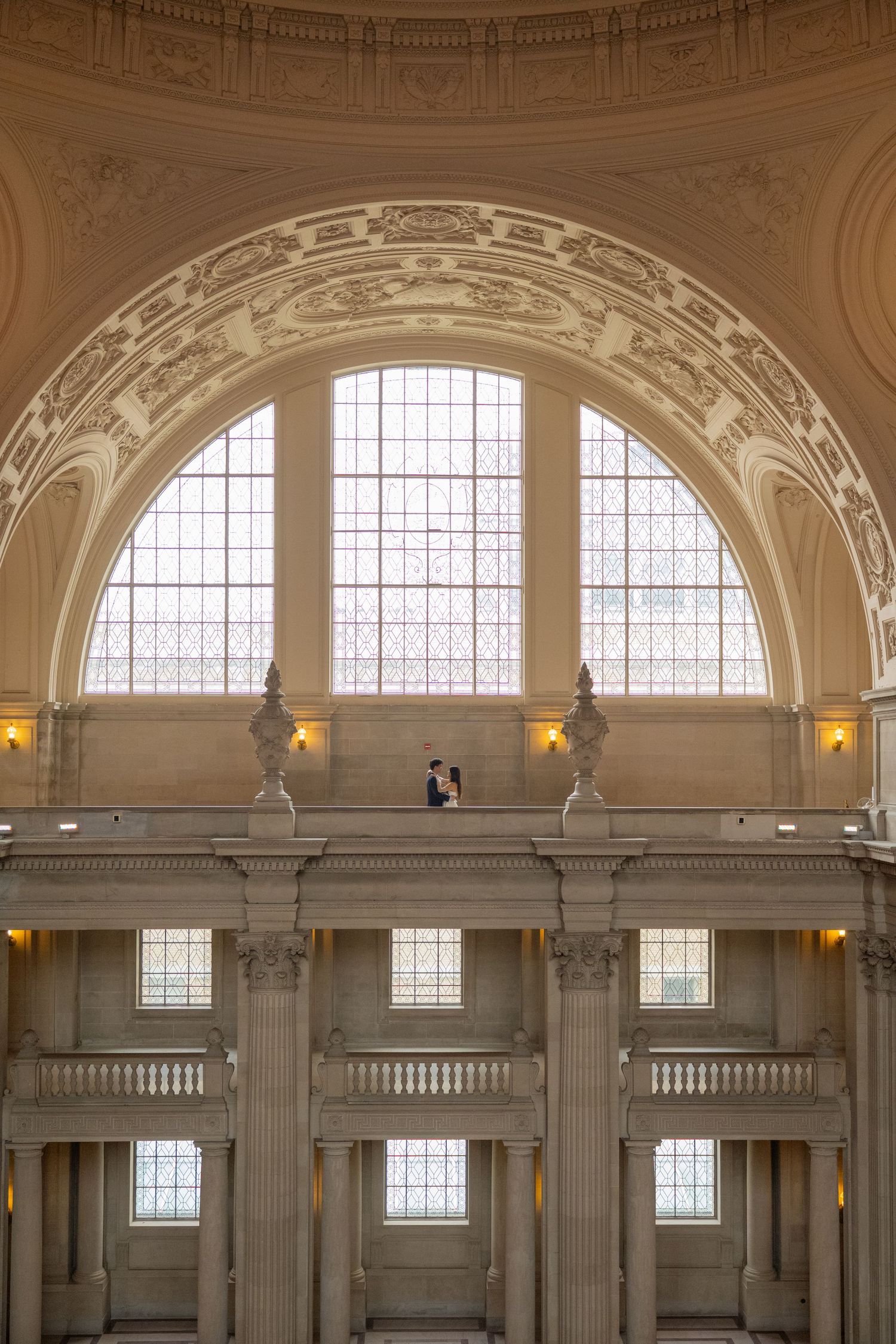 Bride and groom stand on a grand balcony inside San Francisco City Hall beneath an ornate arched window.