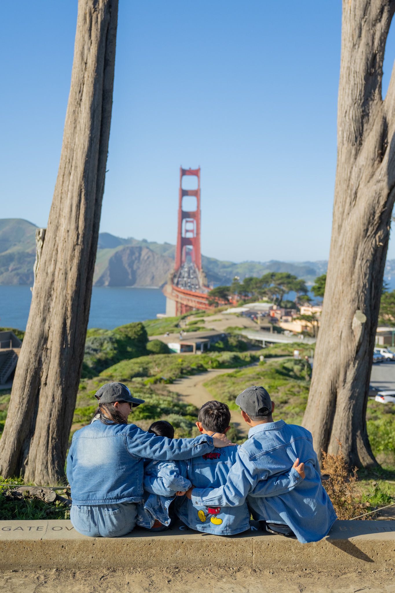 Family in matching denim jackets sits between trees overlooking the iconic Golden Gate Bridge.