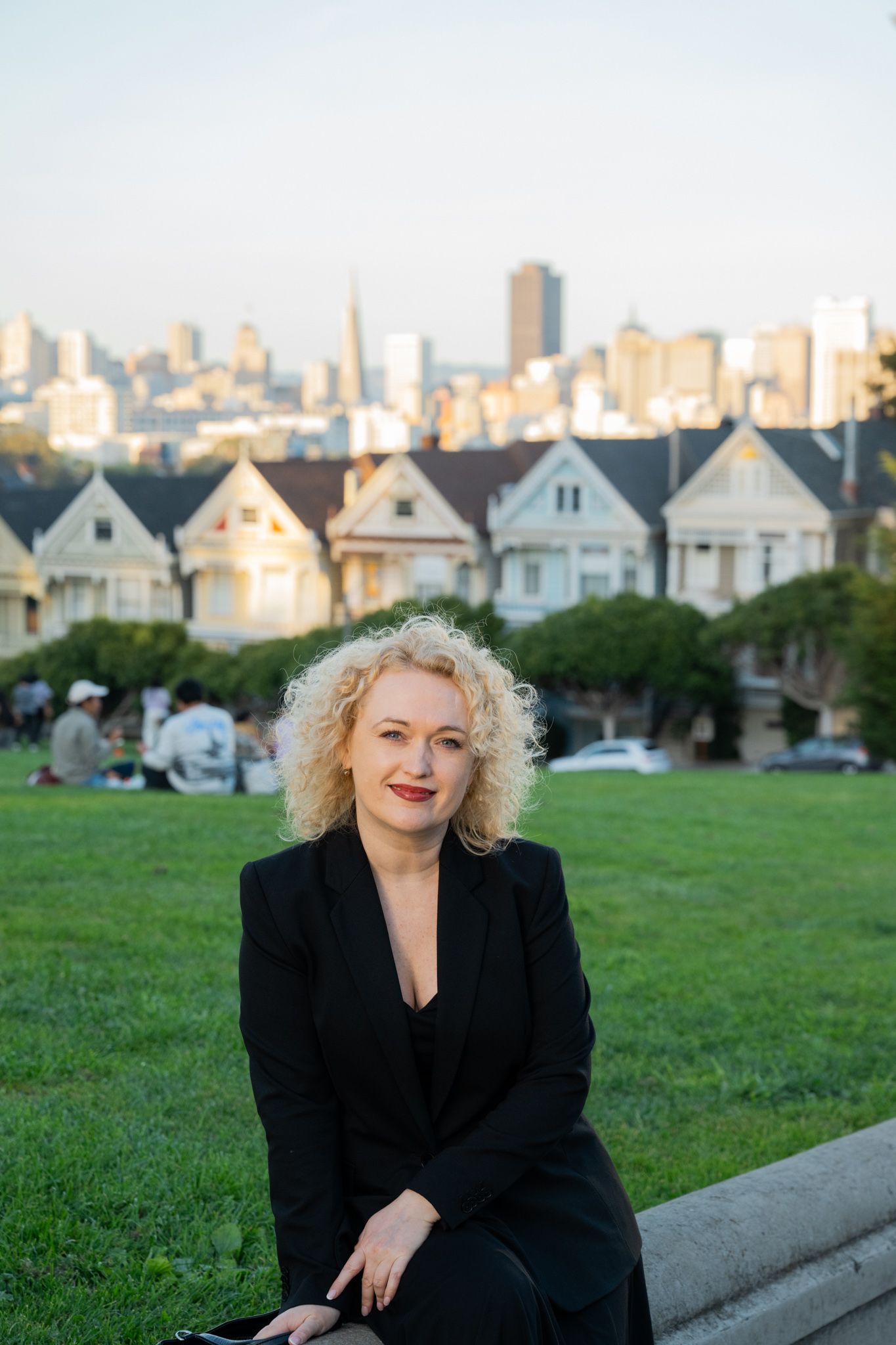 Blonde woman in black blazer sits at Alamo Square Park with Painted Ladies and San Francisco skyline behind her.