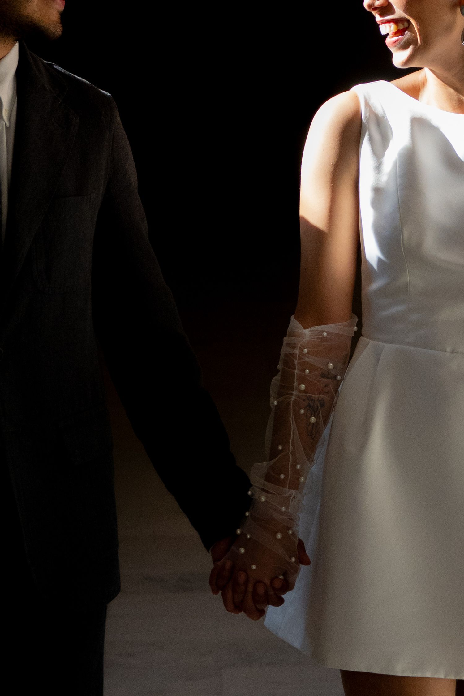 Bride in short white dress with pearl tulle gloves holds grooms hand against dark background.