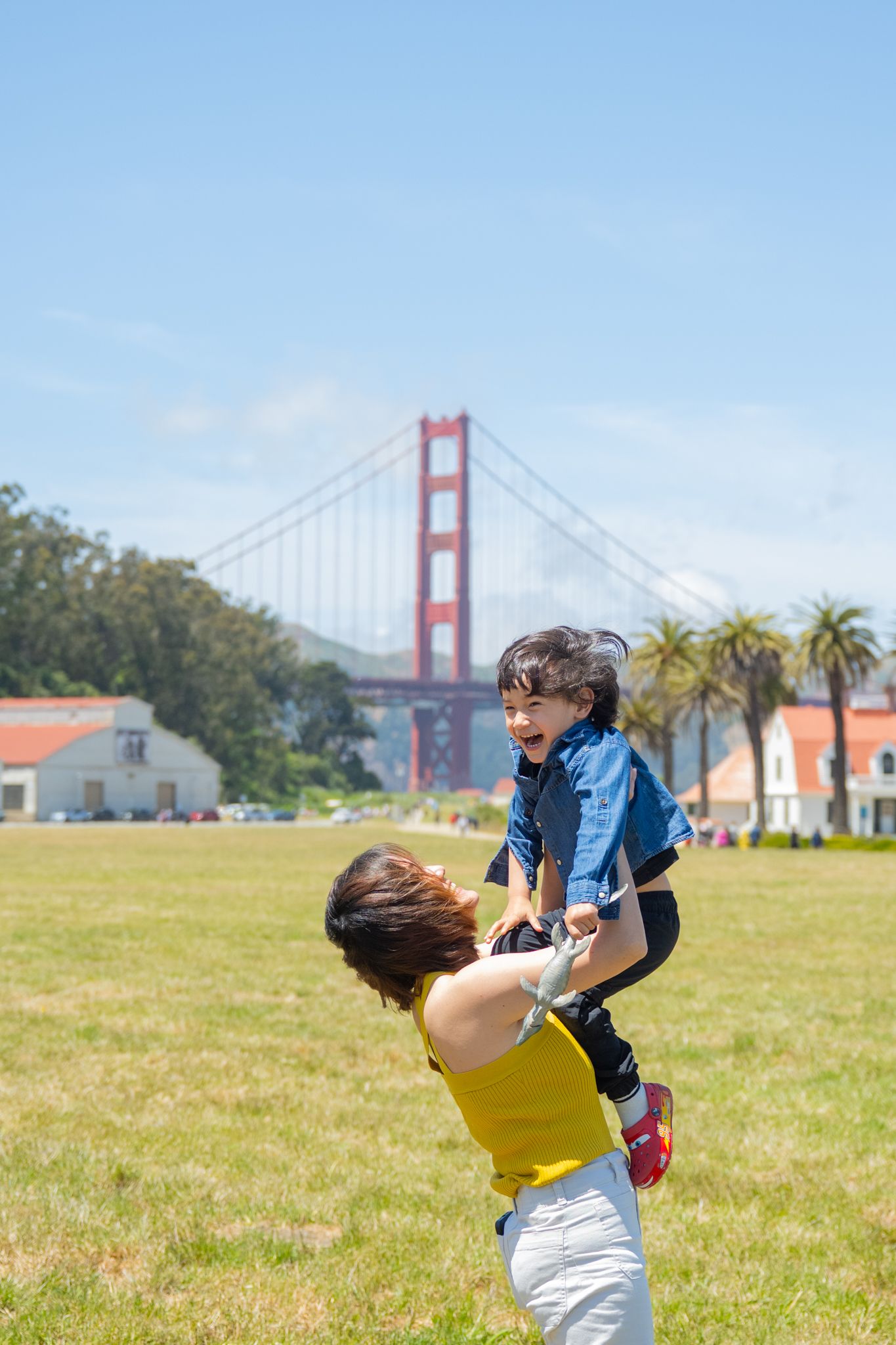 A joyful mother lifts her laughing toddler high in the air at a sunny park near the Golden Gate Bridge.