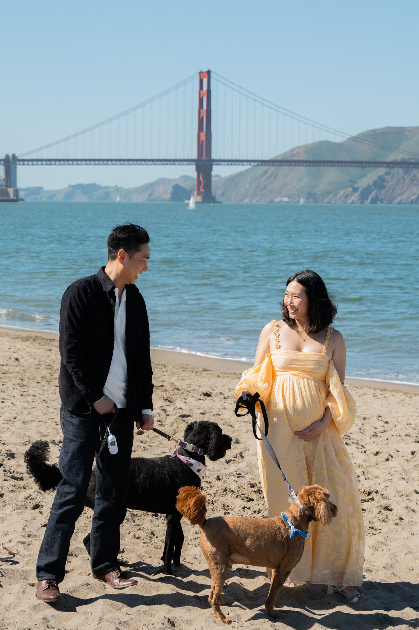 Couple laughing together with three dogs on Baker Beach with the Golden Gate Bridge in the background.