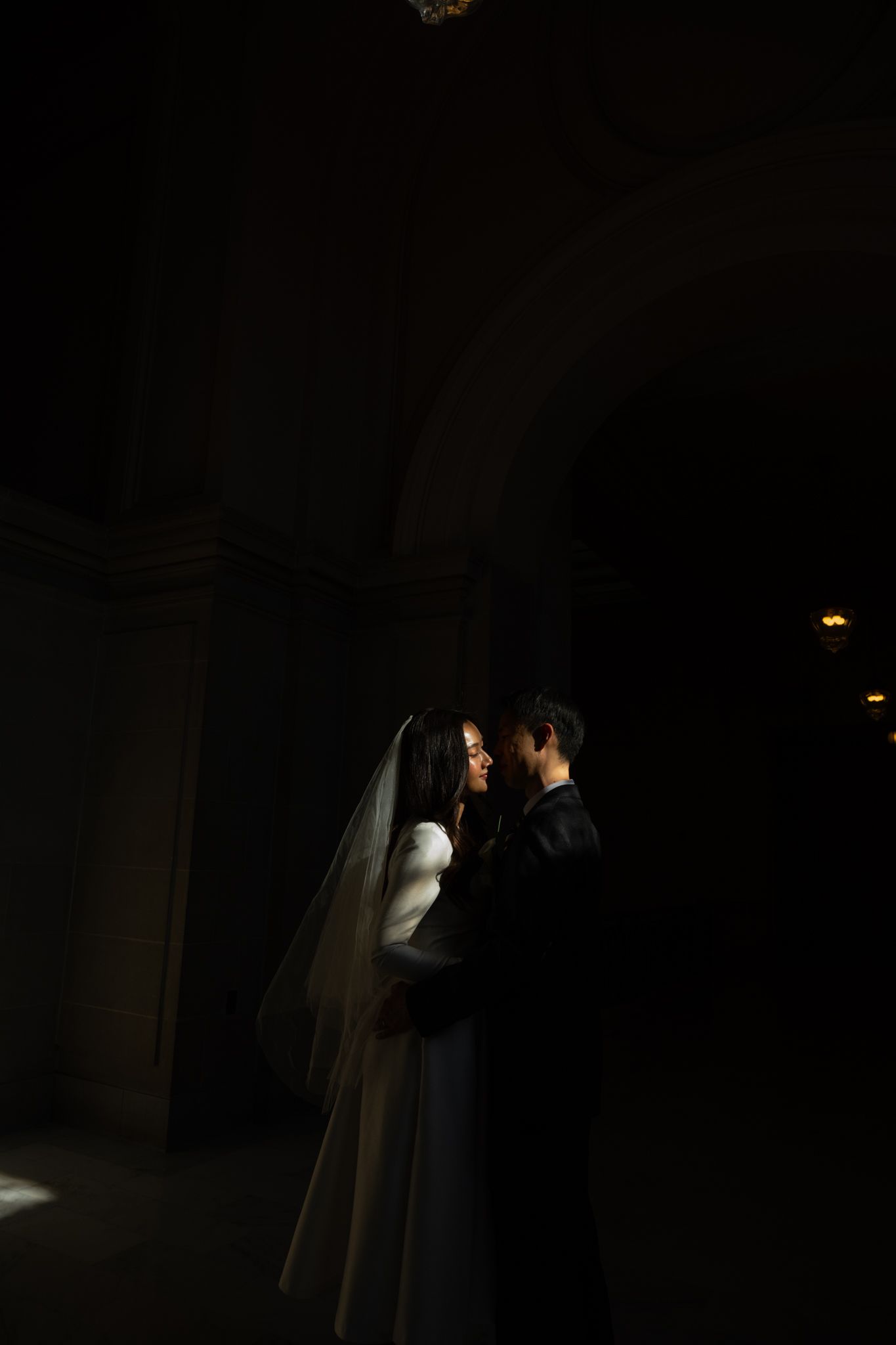 Wedding couple embraces in a darkened architectural space with dramatic light highlighting their silhouettes.