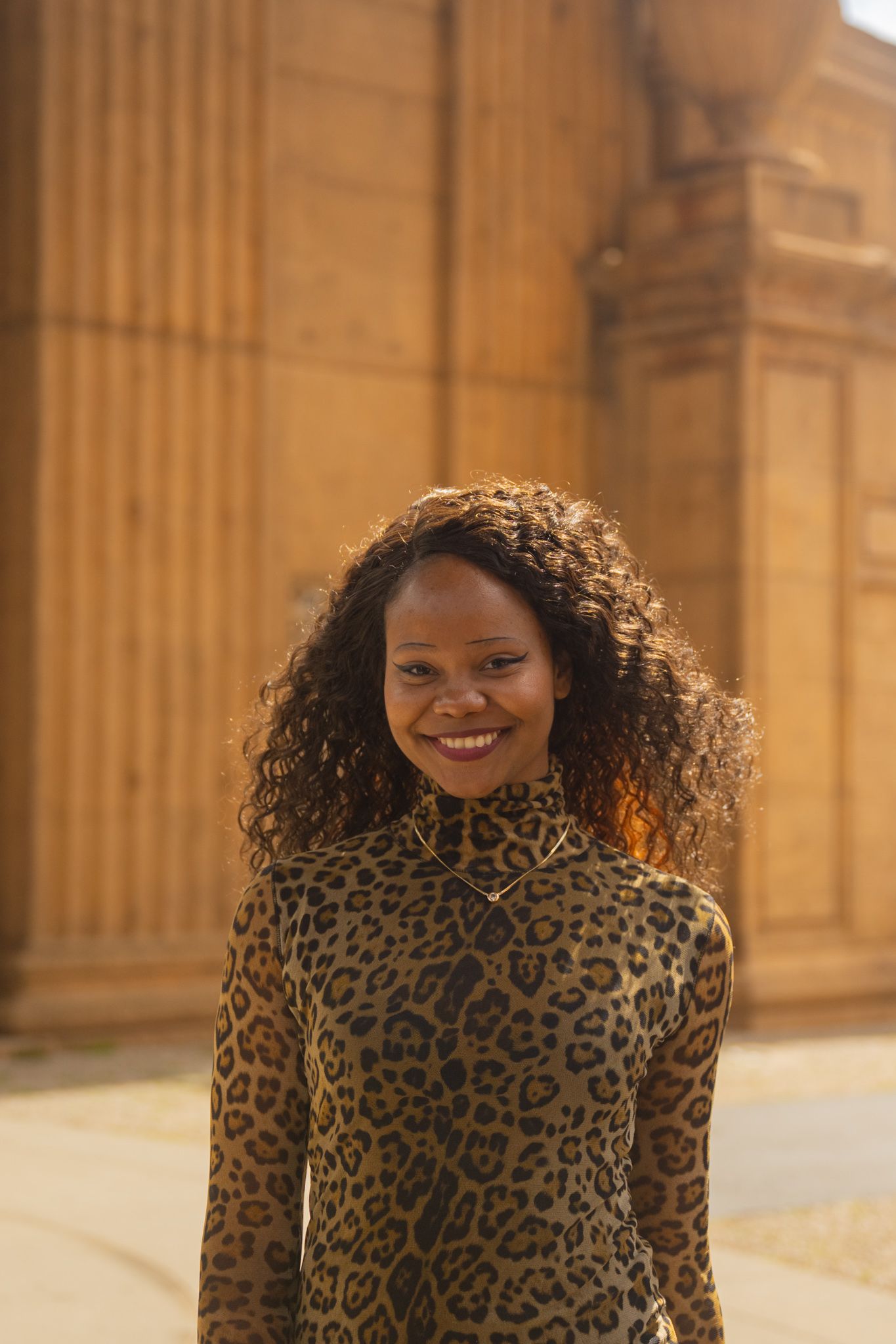 A young Black woman with curly hair smiles confidently in a leopard print top before a golden stone building.