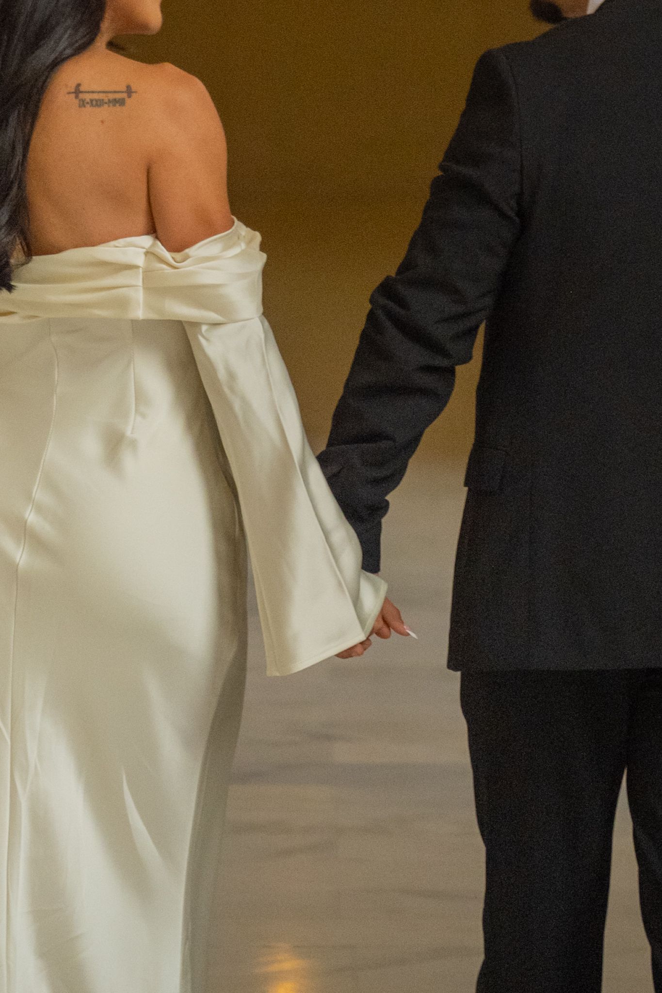 Close-up of bride in off-shoulder white satin gown with bow holding grooms hand at wedding.