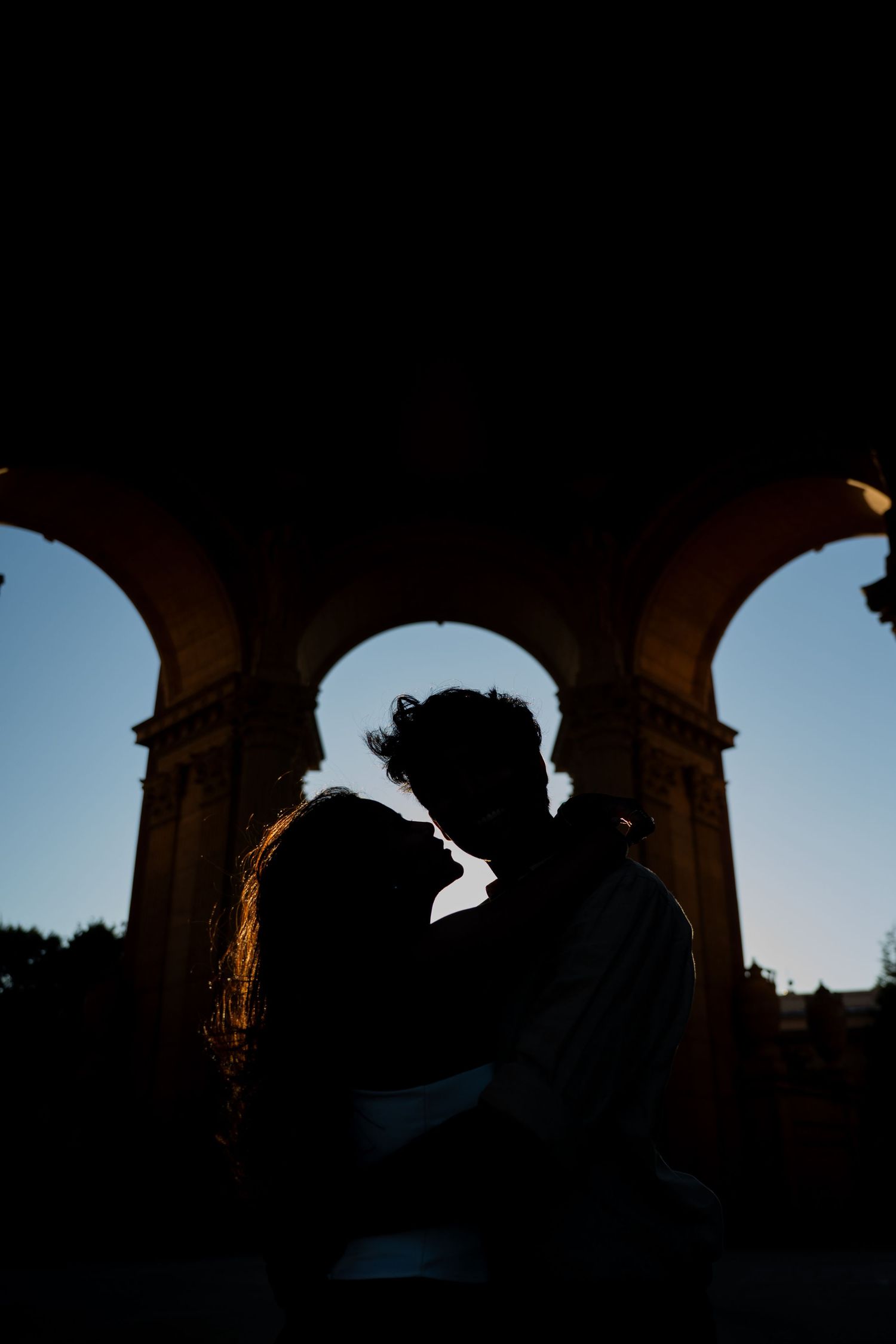 Silhouette of couple nearly kissing framed by elegant stone arches at dusk with blue sky backdrop.