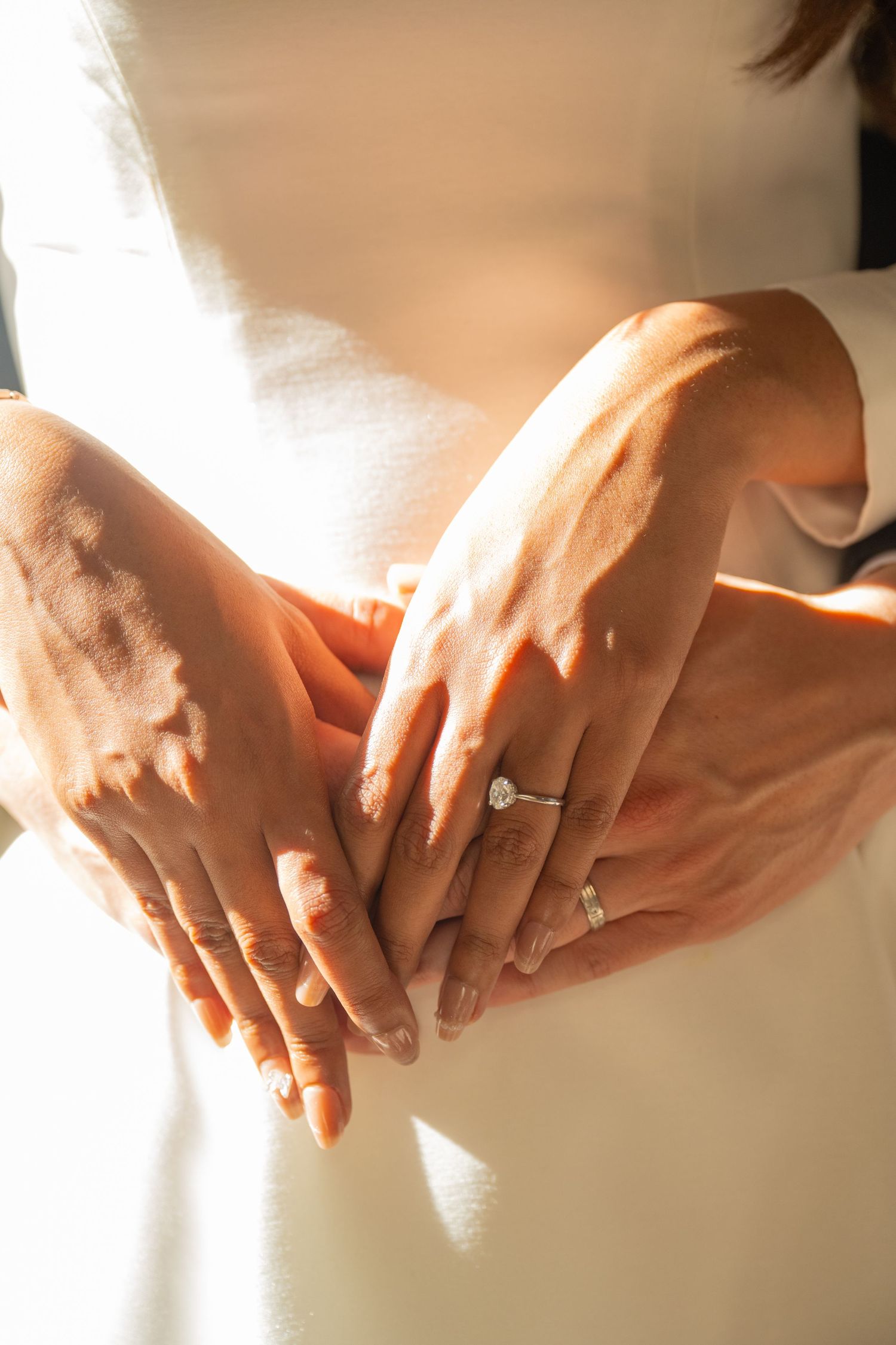 Close-up of bride and grooms hands together showing diamond engagement ring in warm sunlight.