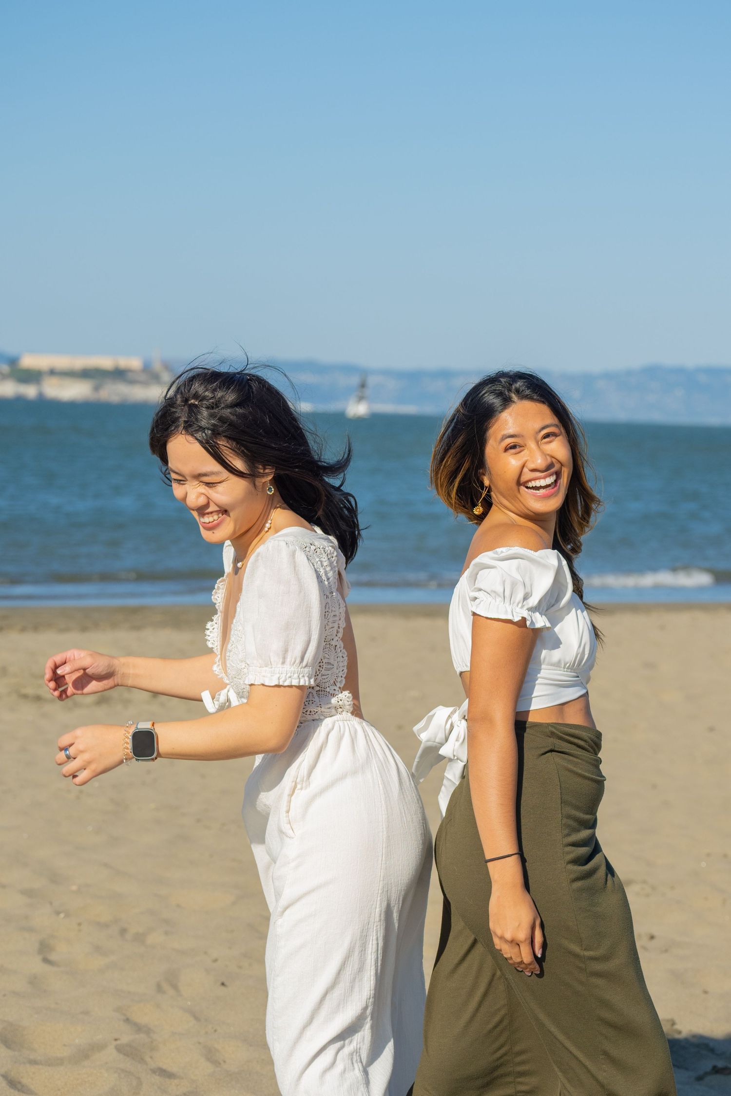 Two joyful young Asian women laugh and walk together on a sunny sandy beach.