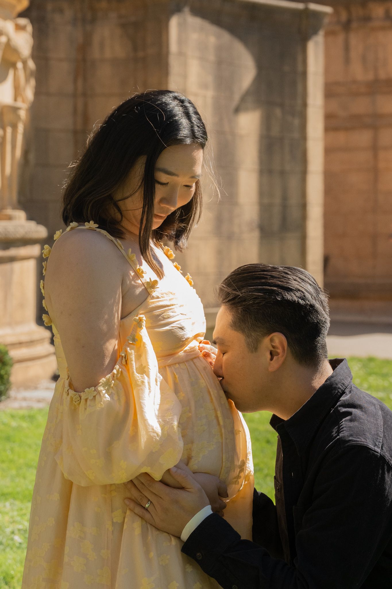 A man tenderly kisses a pregnant woman's belly outdoors near stone columns, as she looks down lovingly in a yellow dress.