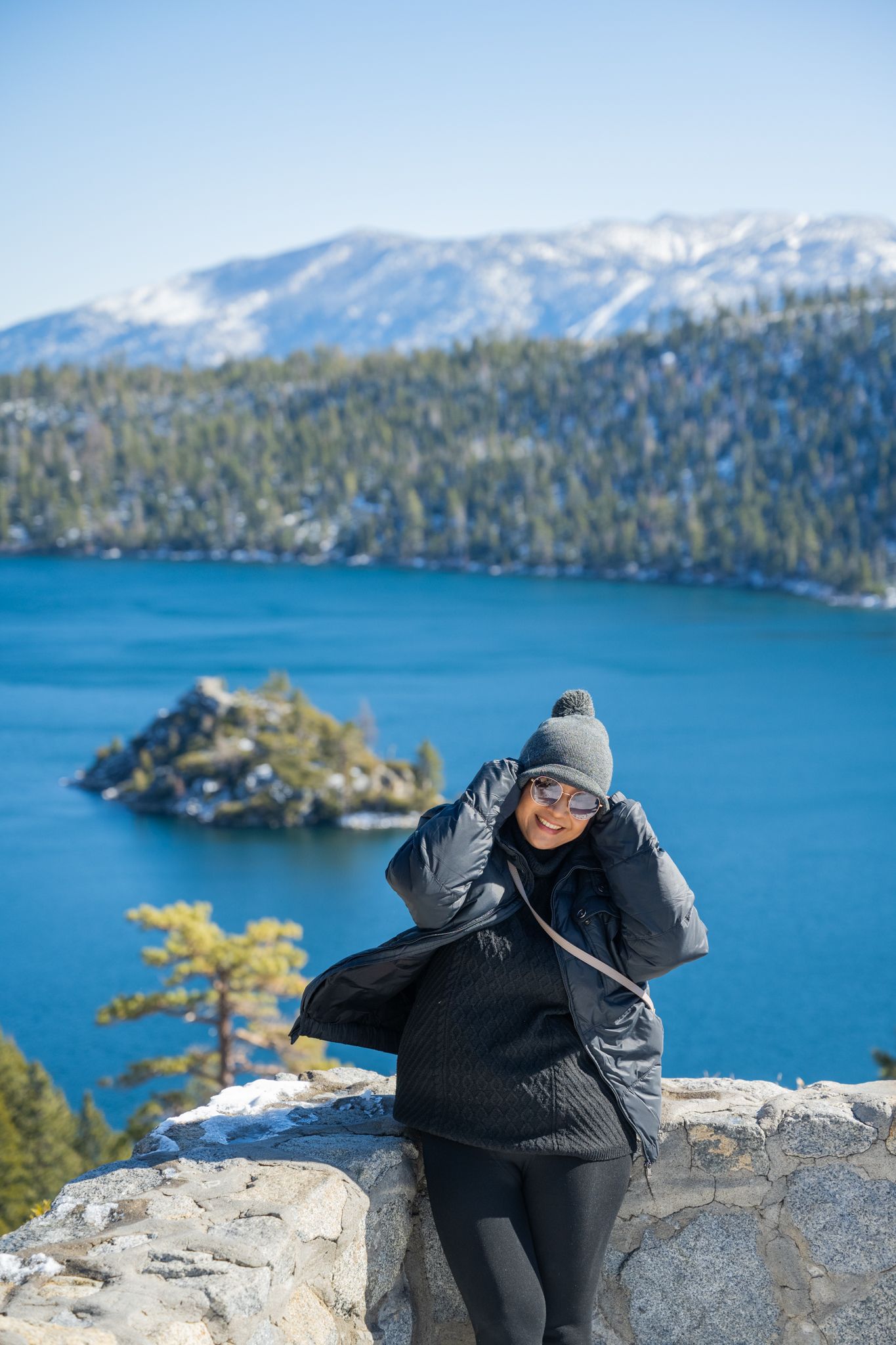 A woman in winter gear smiles at Emerald Bay, Lake Tahoe, with snow-capped mountains and blue water behind her.