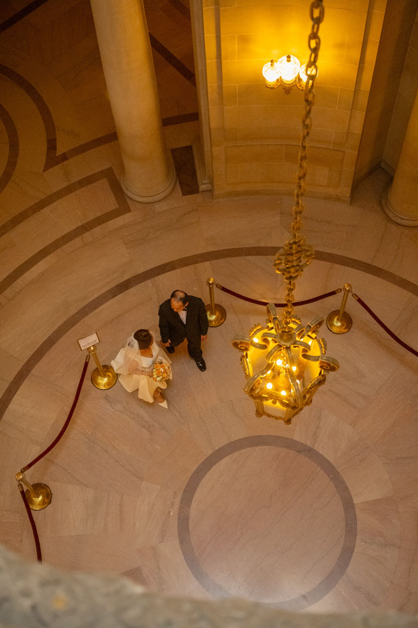 Aerial view of bride and groom standing in ornate marble rotunda with golden chandelier at San Francisco City Hall.
