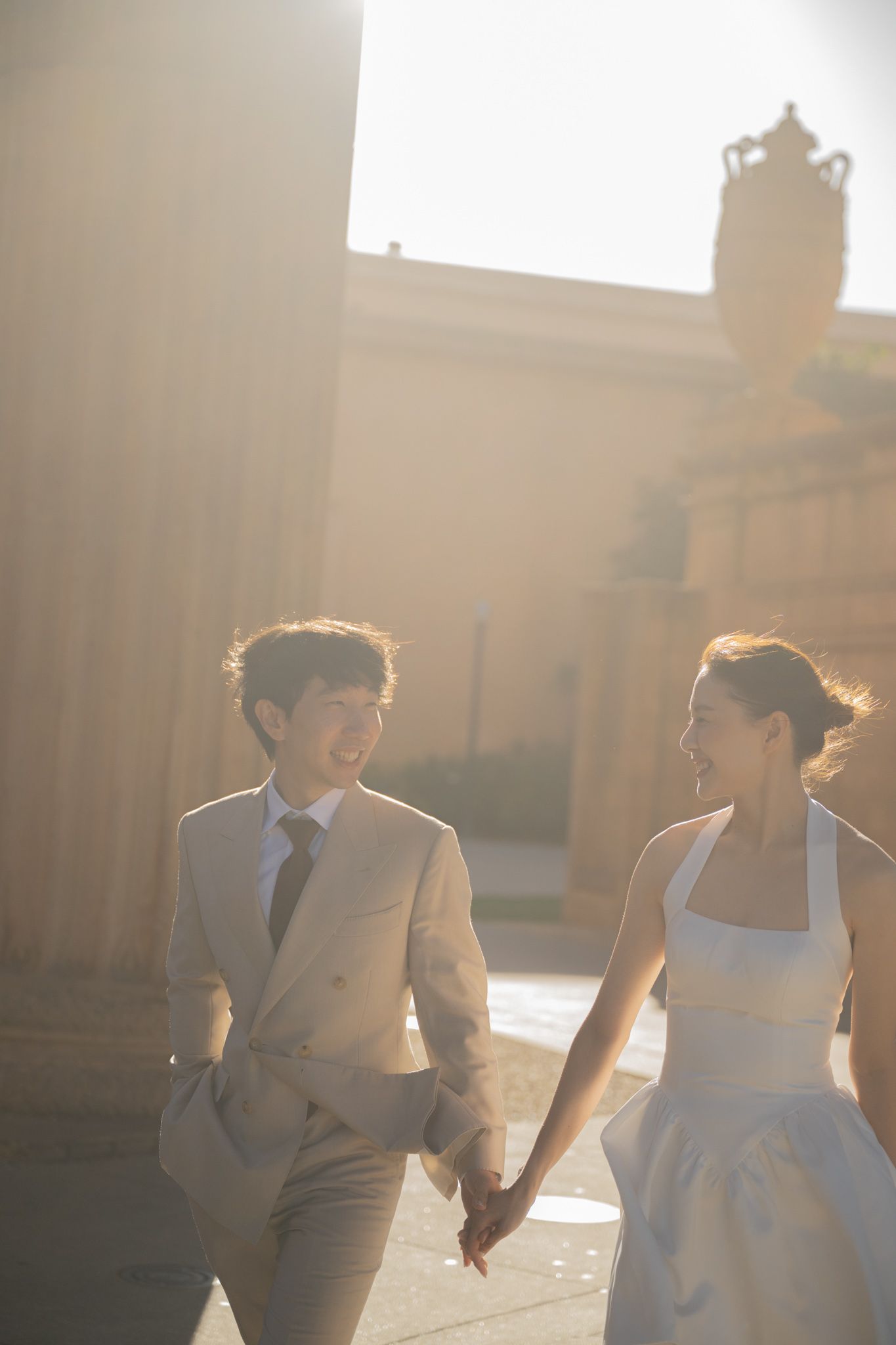 Smiling newlyweds holding hands and walking past grand columns in warm sunlight during their wedding day.
