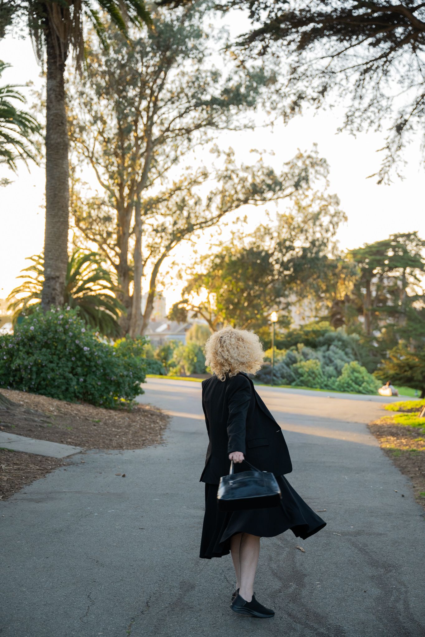 Curly blonde woman in black coat and handbag walks along a sunlit park path at golden hour.