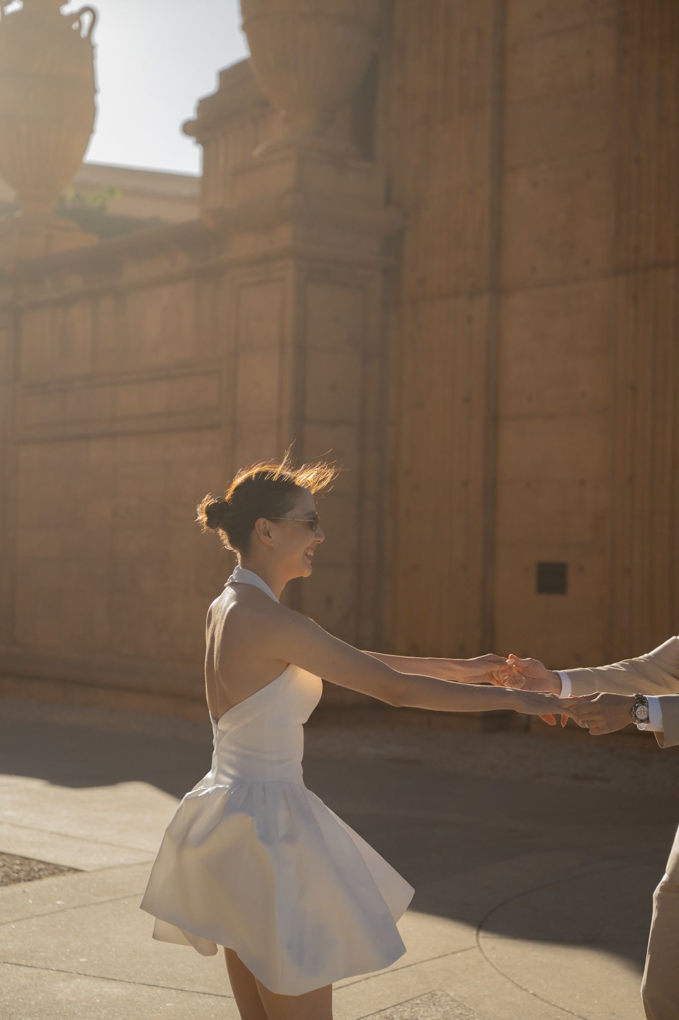 A bride in a white mini dress holds hands with her groom in golden sunlight outside a historic stone building.