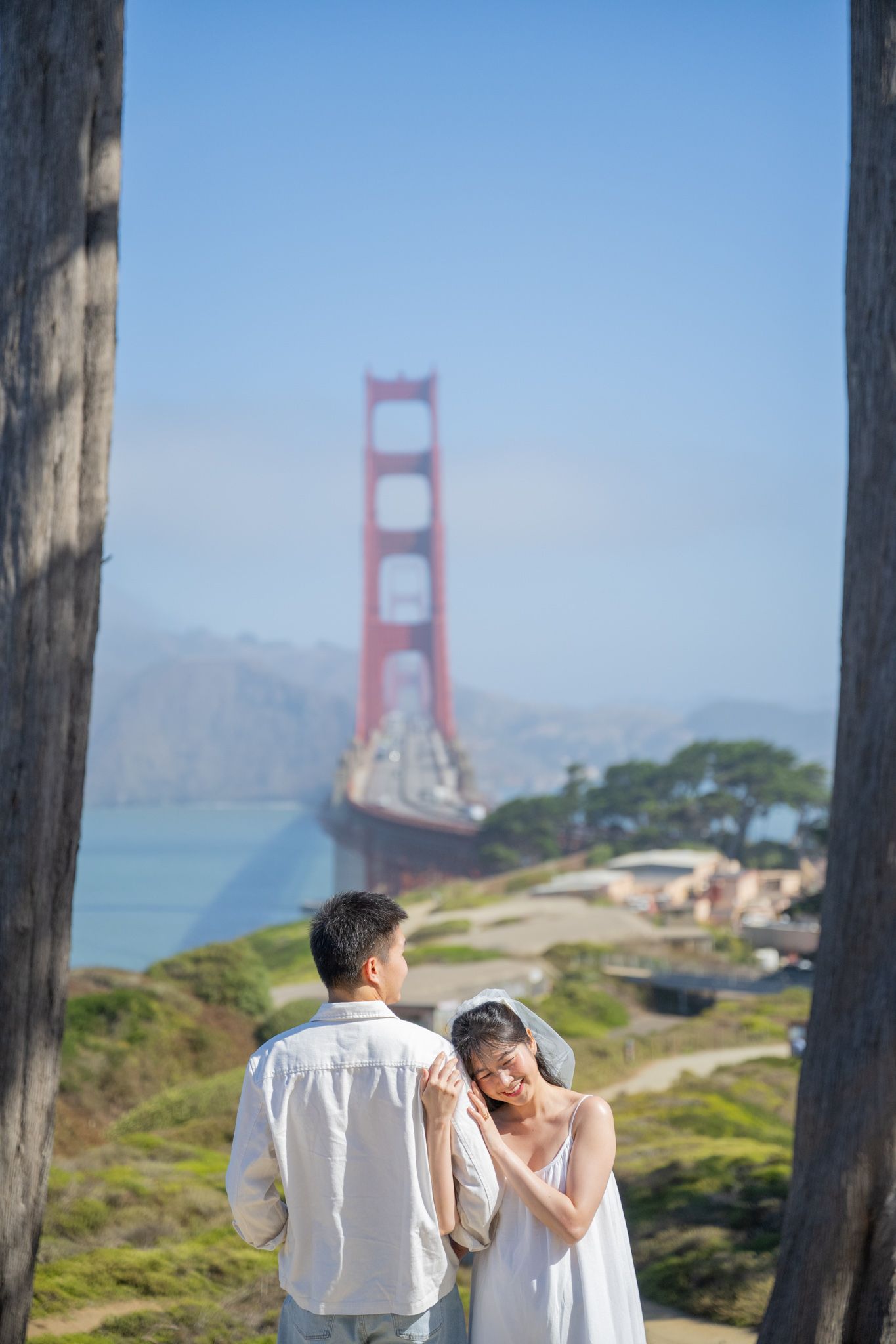 Couple in white attire poses romantically before the iconic Golden Gate Bridge on a sunny day in San Francisco.