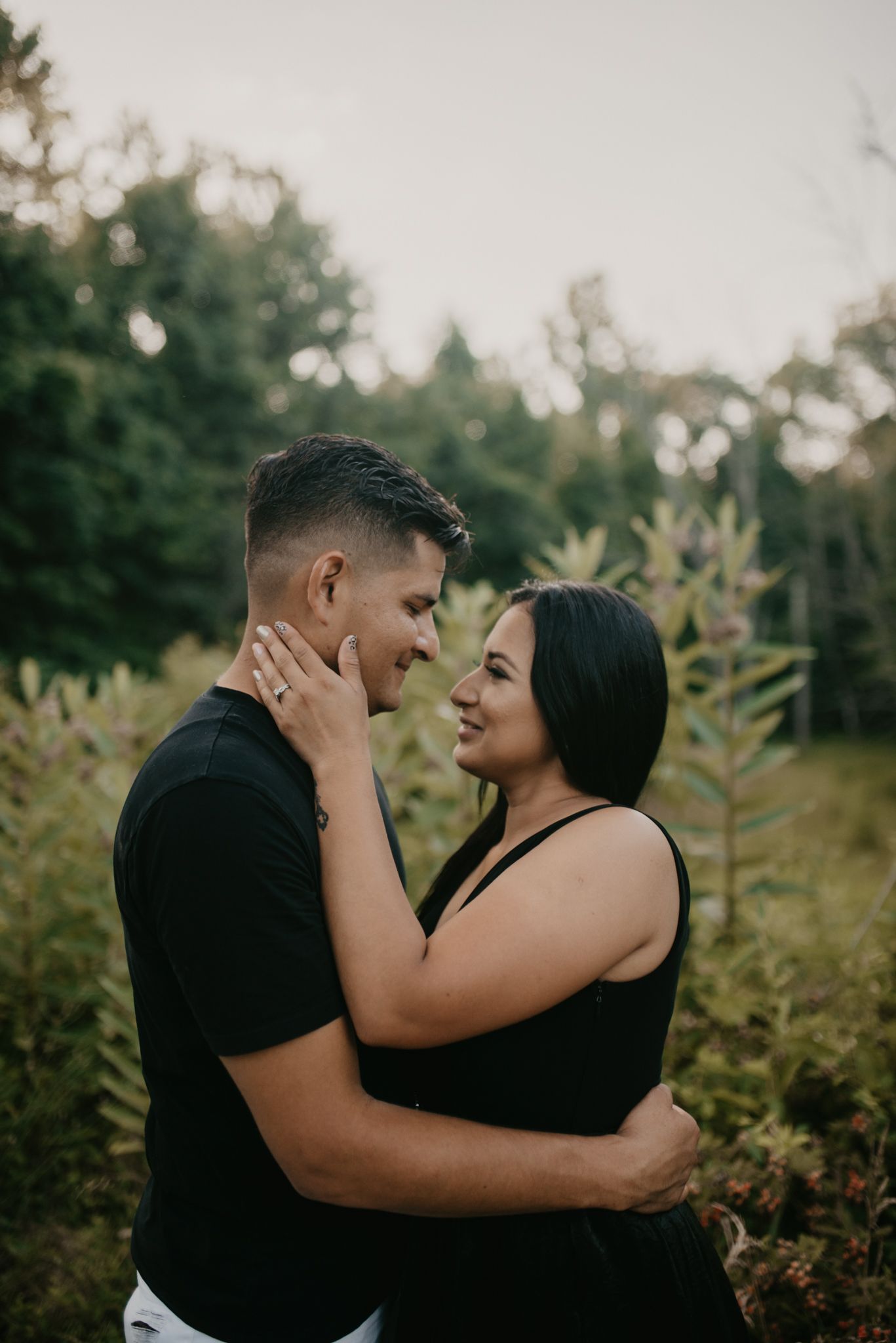 A romantic couple embraces in a natural outdoor setting with green foliage at sunset.