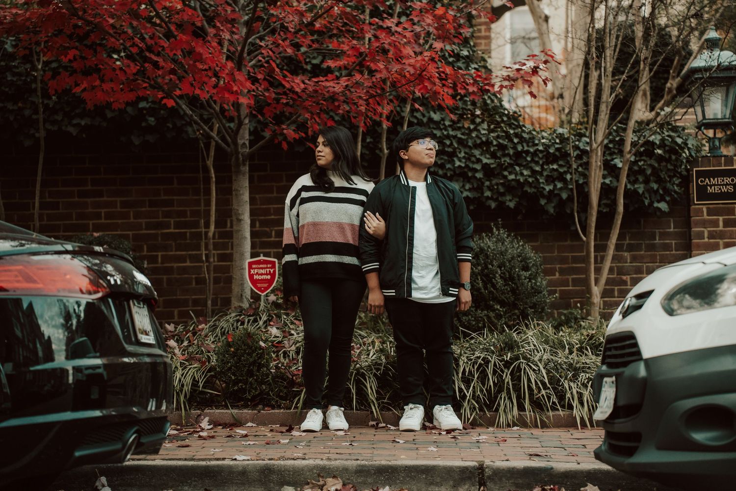 Two people stand on a sidewalk between parked cars with red autumn foliage visible in the background in Old town Alexandria