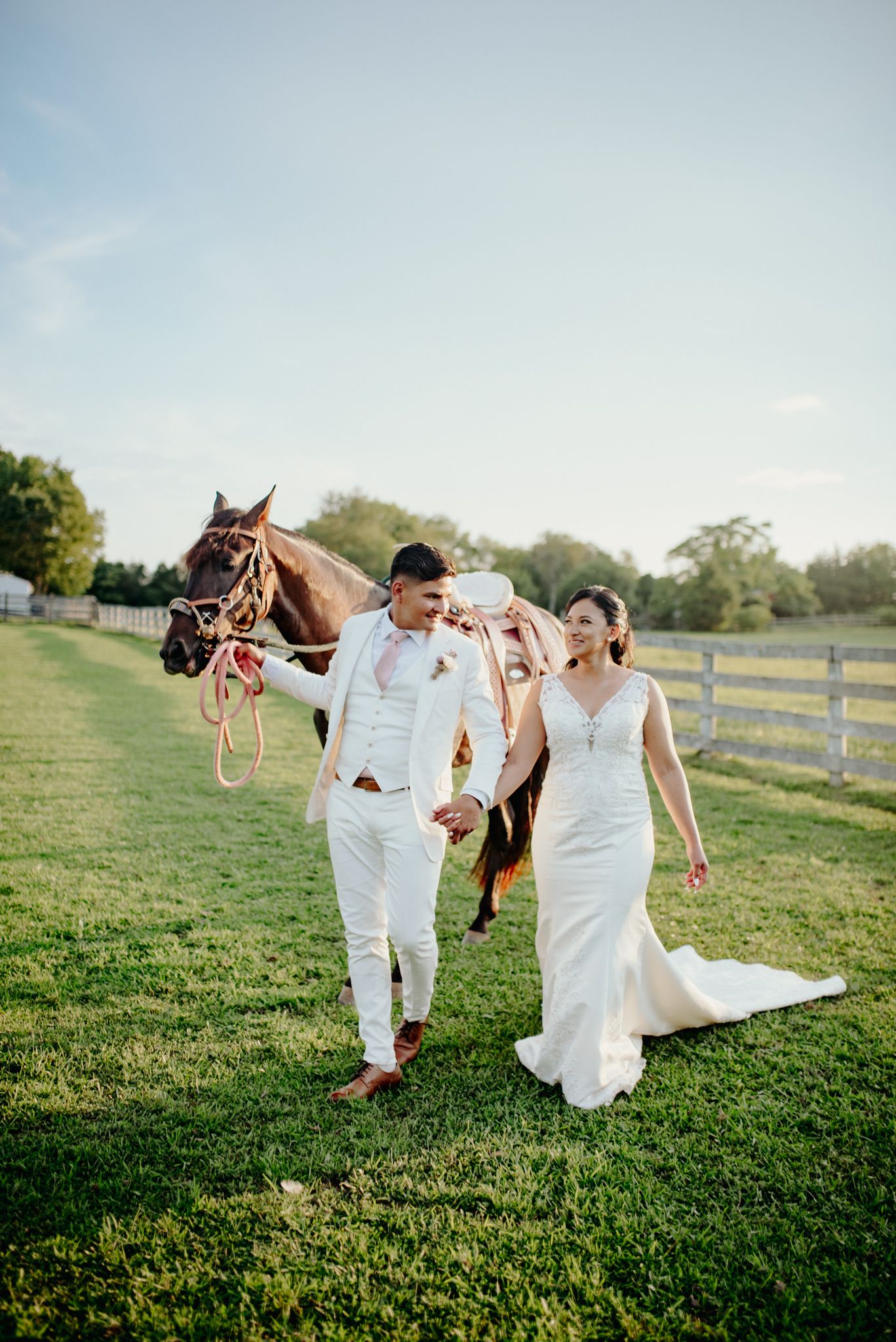 A couple in white wedding attire playfully runs and dances together on a grassy hillside during golden hour.