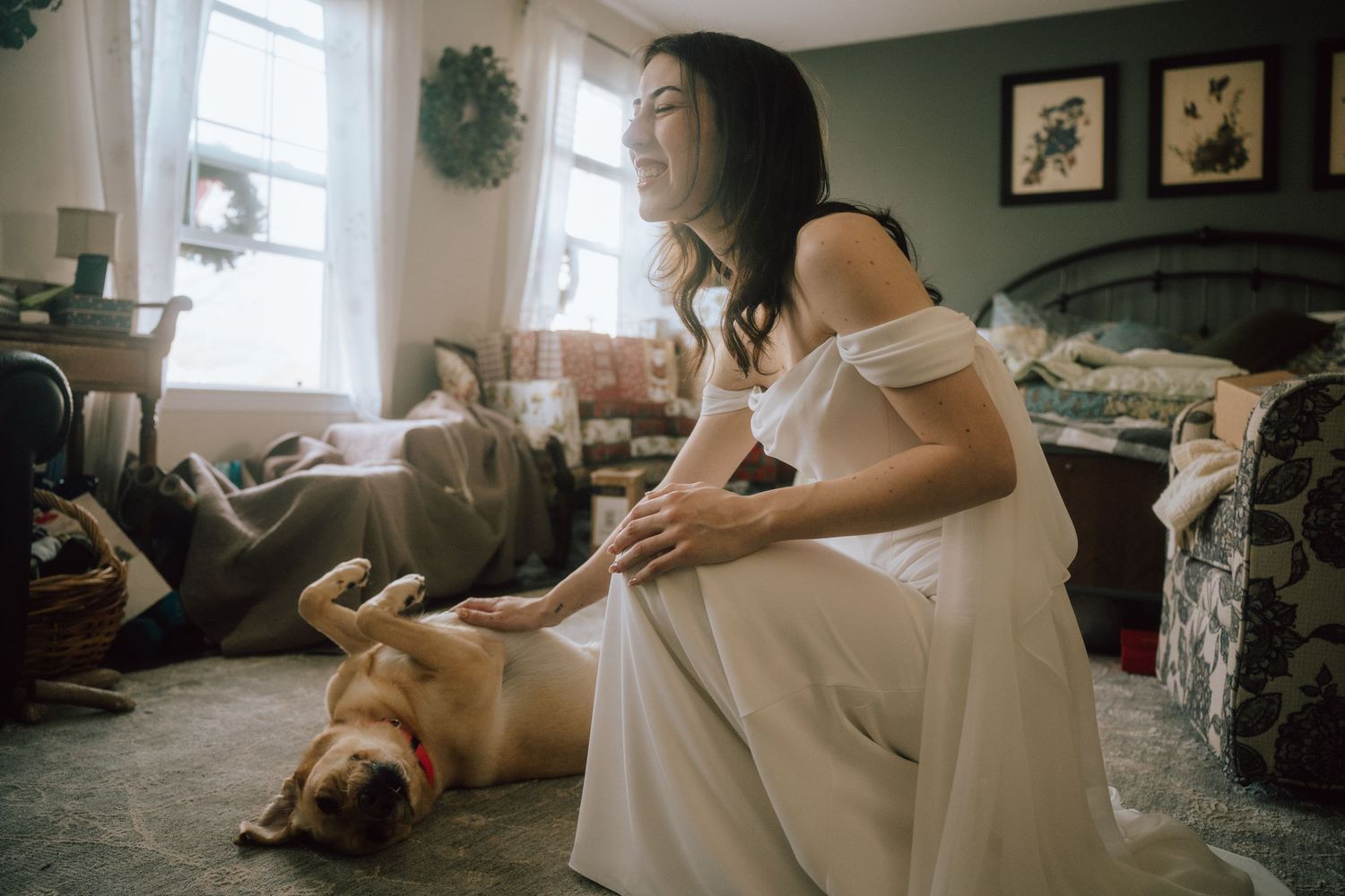 Someone in a white dress sits on the floor petting a relaxed golden retriever dog lying on its back in a cozy living room.
