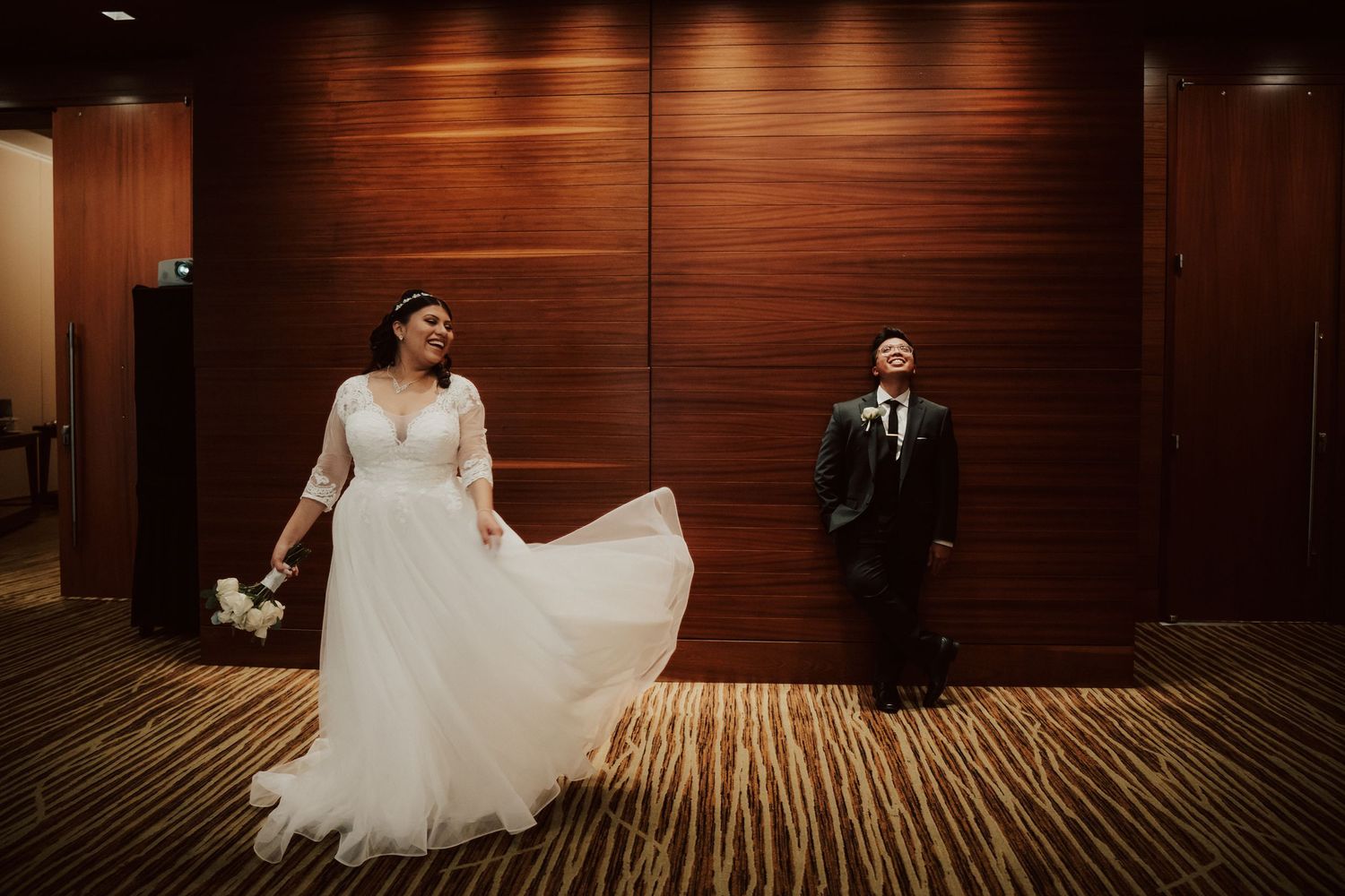 A bride in a white wedding gown sits gracefully on a wooden bench against a dark paneled wall.